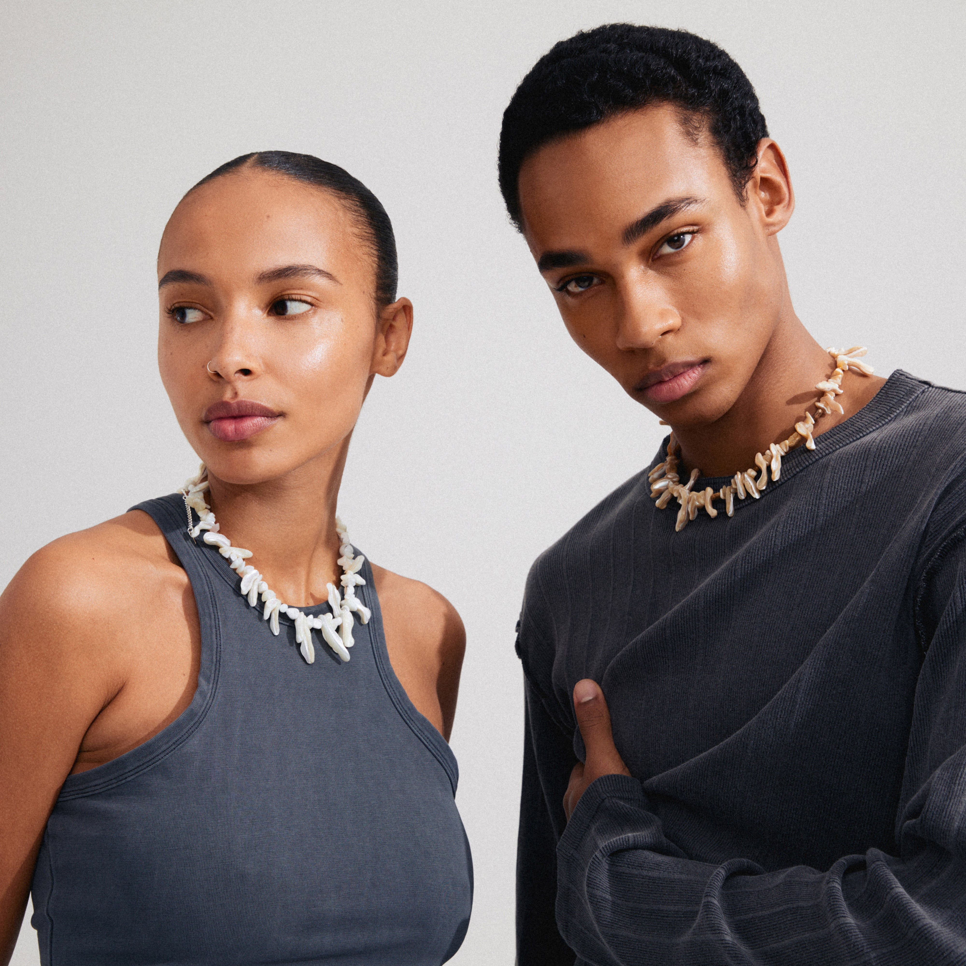 Two young adults wear Pilgrim's LIGHT seashell necklace, featuring chunky, irregular mother-of-pearl beads, showcasing a bohemian summer vibe against a plain background.
