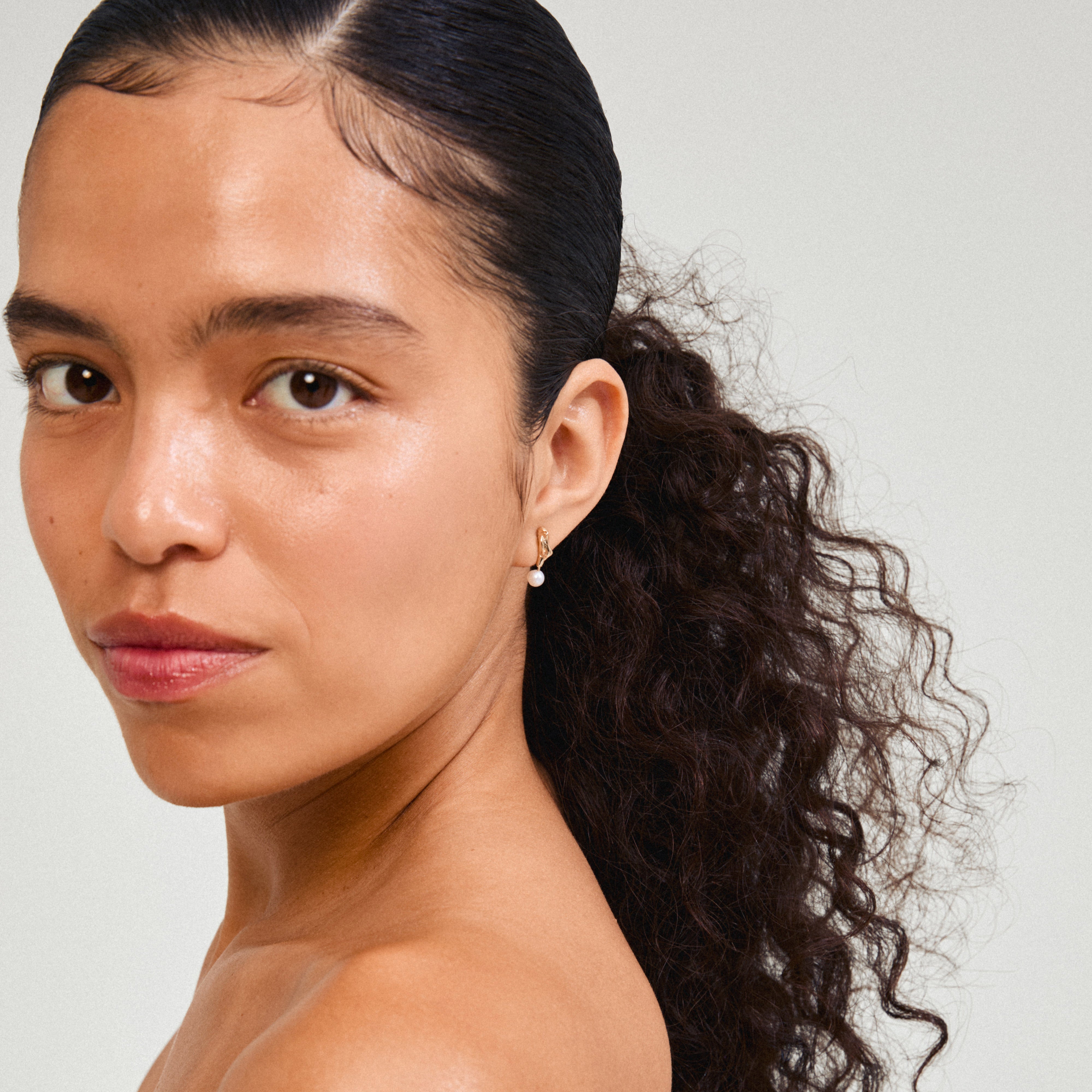 Close-up of STARLIGHT pearl earrings in gold-plated design, featuring organic wavy shapes and genuine freshwater pearl pendants, worn by a woman with long hair.