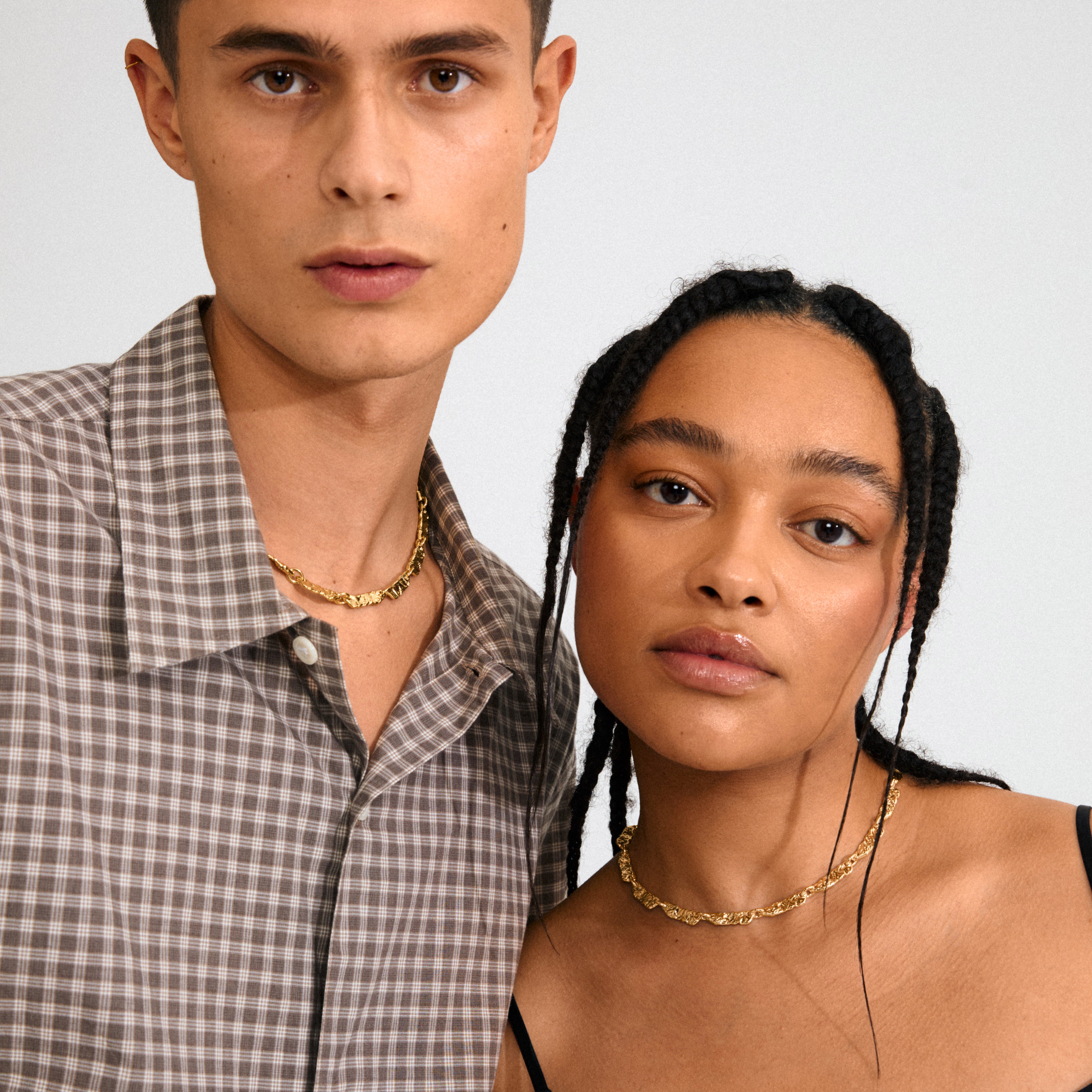 Young man and woman wear Pilgrim's INSTANT gold-plated necklaces with textured links, showcasing stylish, handcrafted elegance against a simple backdrop.