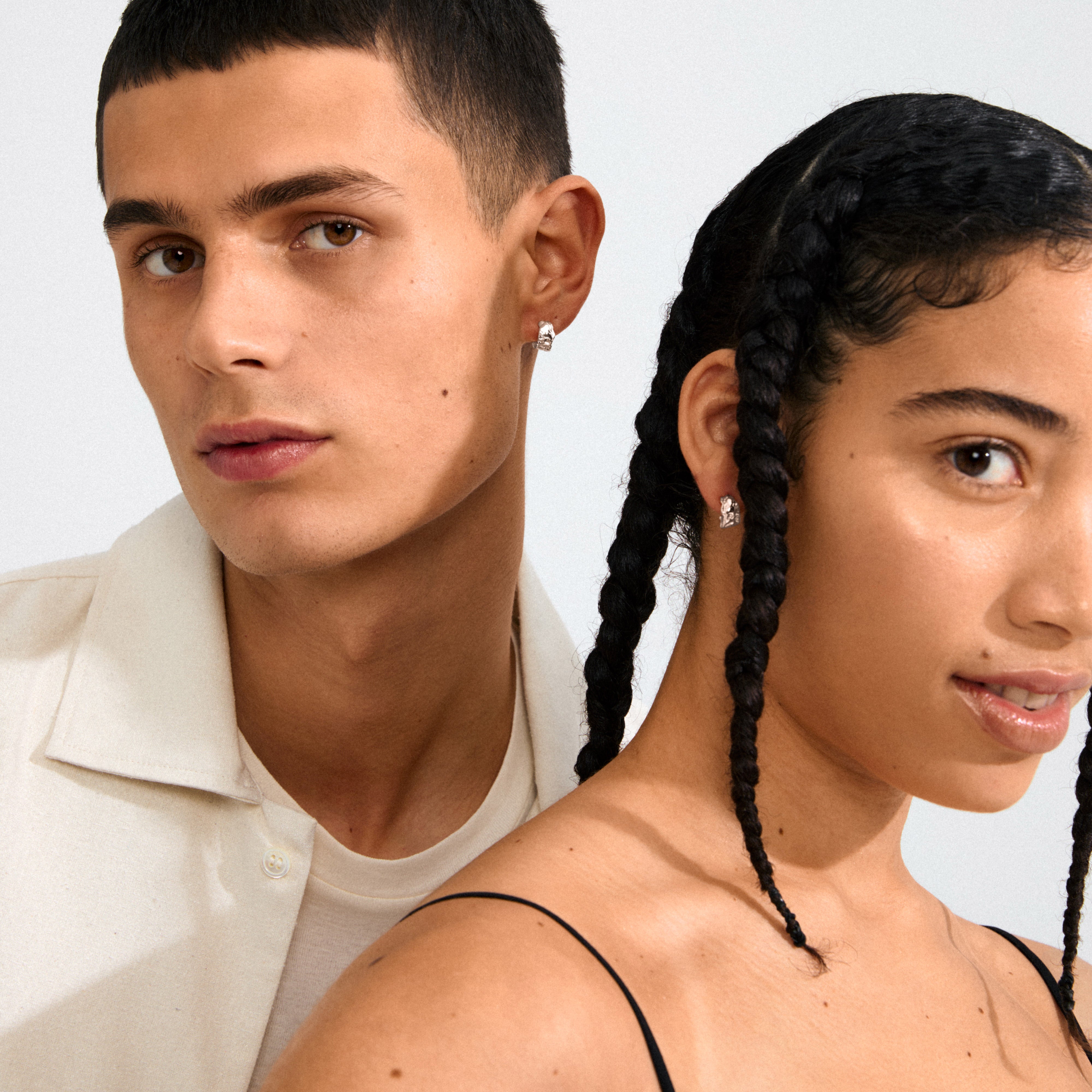 Two young adults wear Pilgrim's INSTANT silver-plated huggie hoops, showcasing their timeless elegance and organic texture in a stylish portrait.