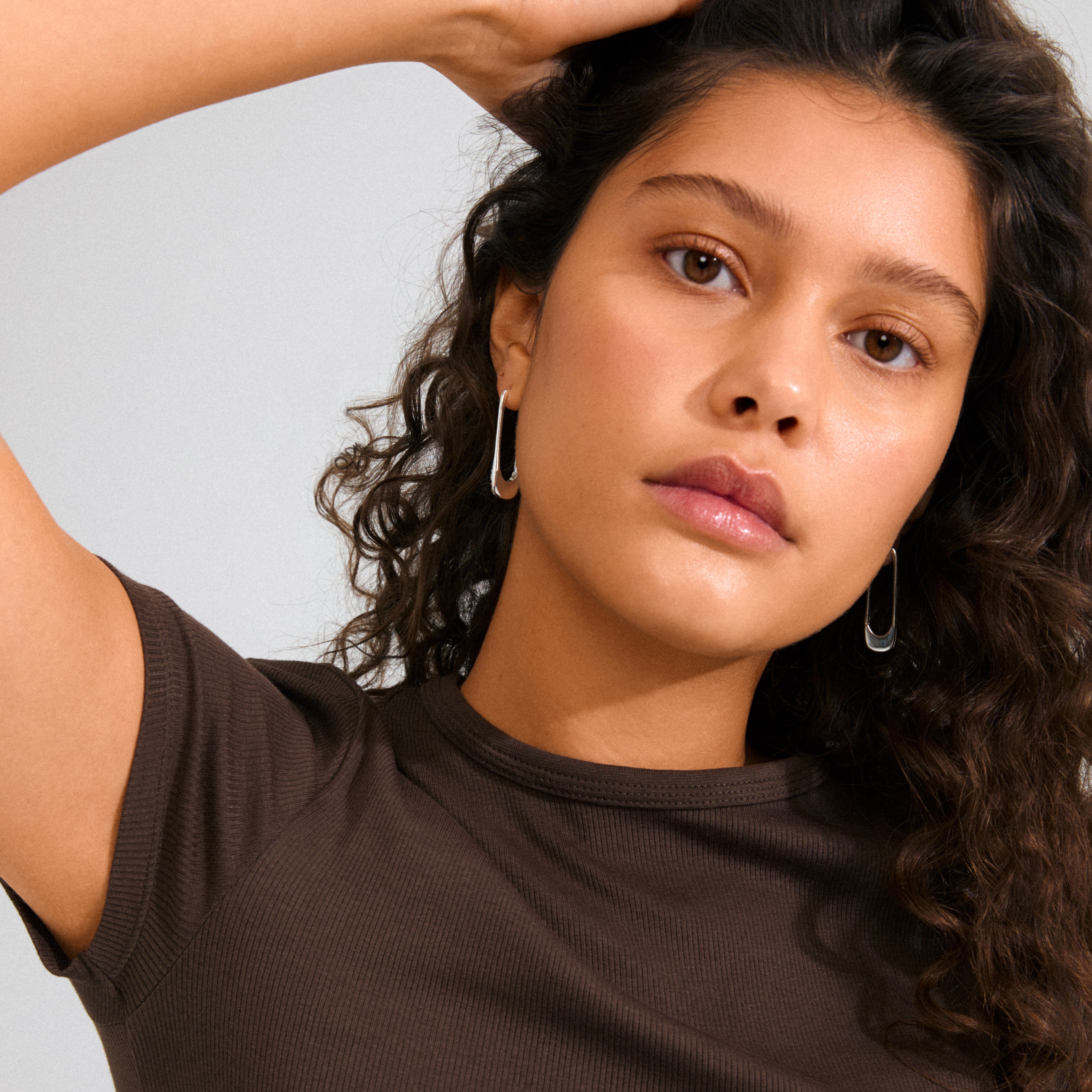 Young woman wearing Pilgrim's INSTANT hoops, silver-plated earrings inspired by safety pins, with Preciosa crystals, styled with curly hair and a brown top.