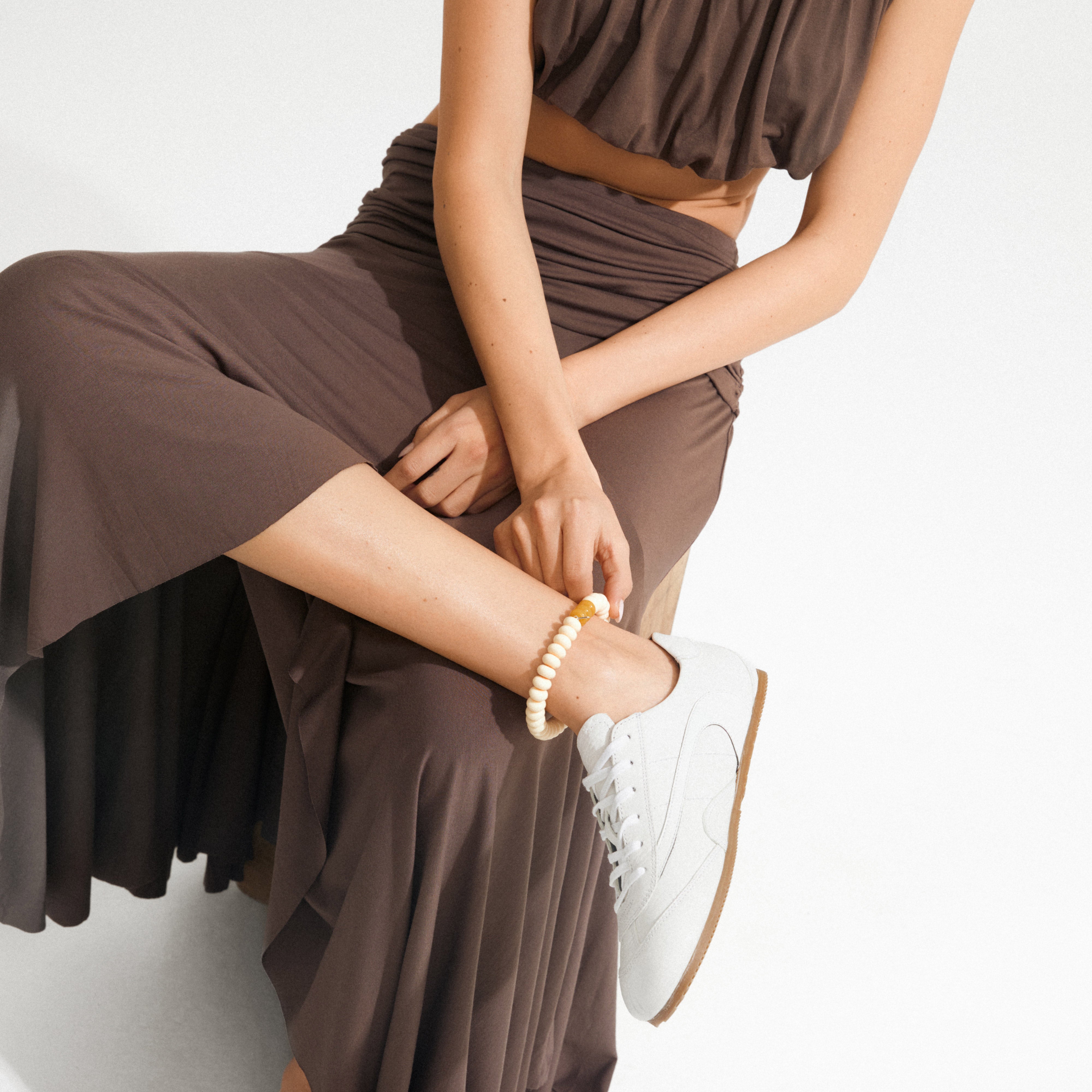 Model adjusting ECLIPSE anklet with chunky ivory beads on bare leg, paired with a chocolate-brown dress and white sneakers.