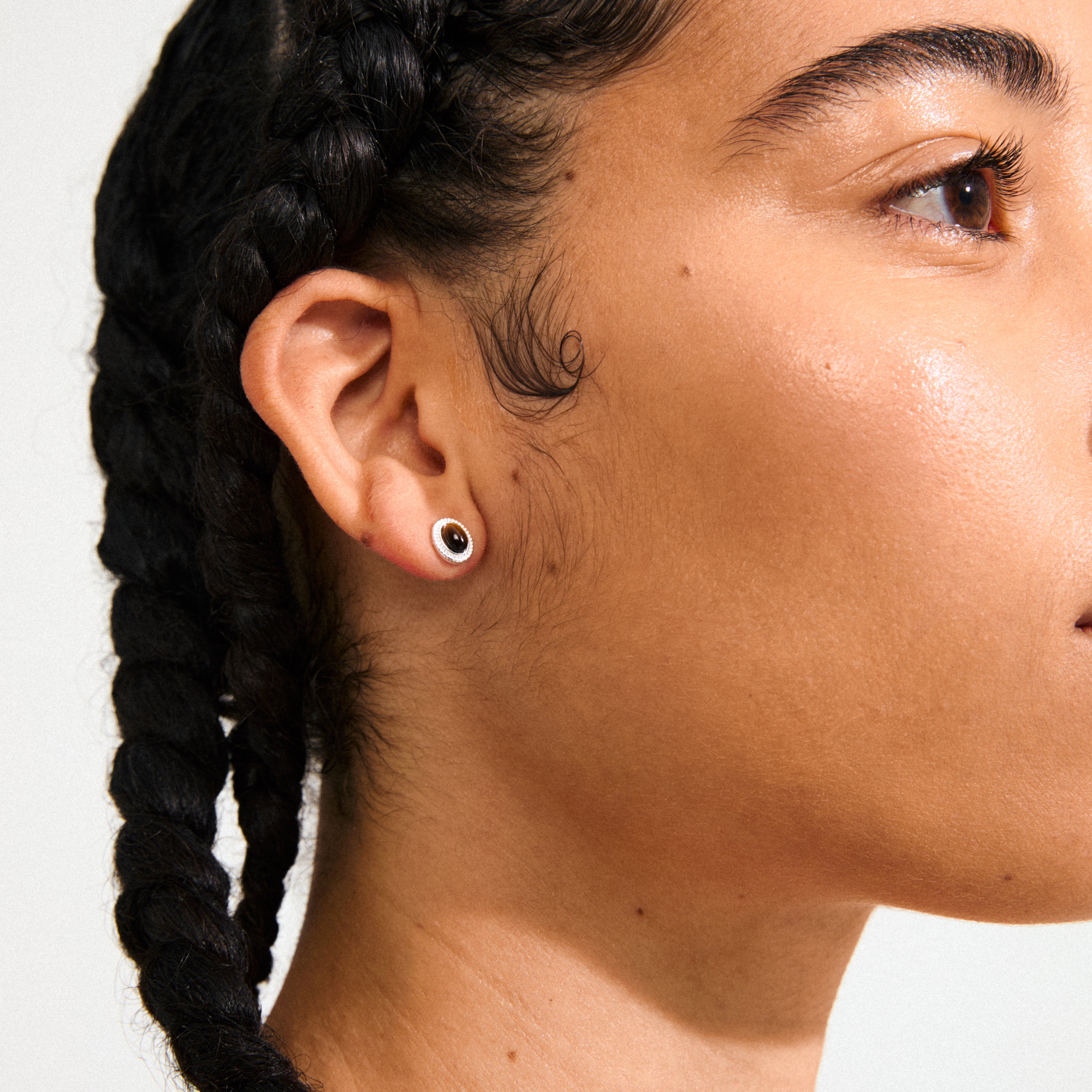 Close-up of a person's ear showcasing Pilgrim's BETWEEN silver-plated stud earrings, featuring cabochon-cut Tiger Eye stones in vintage-inspired gold settings.