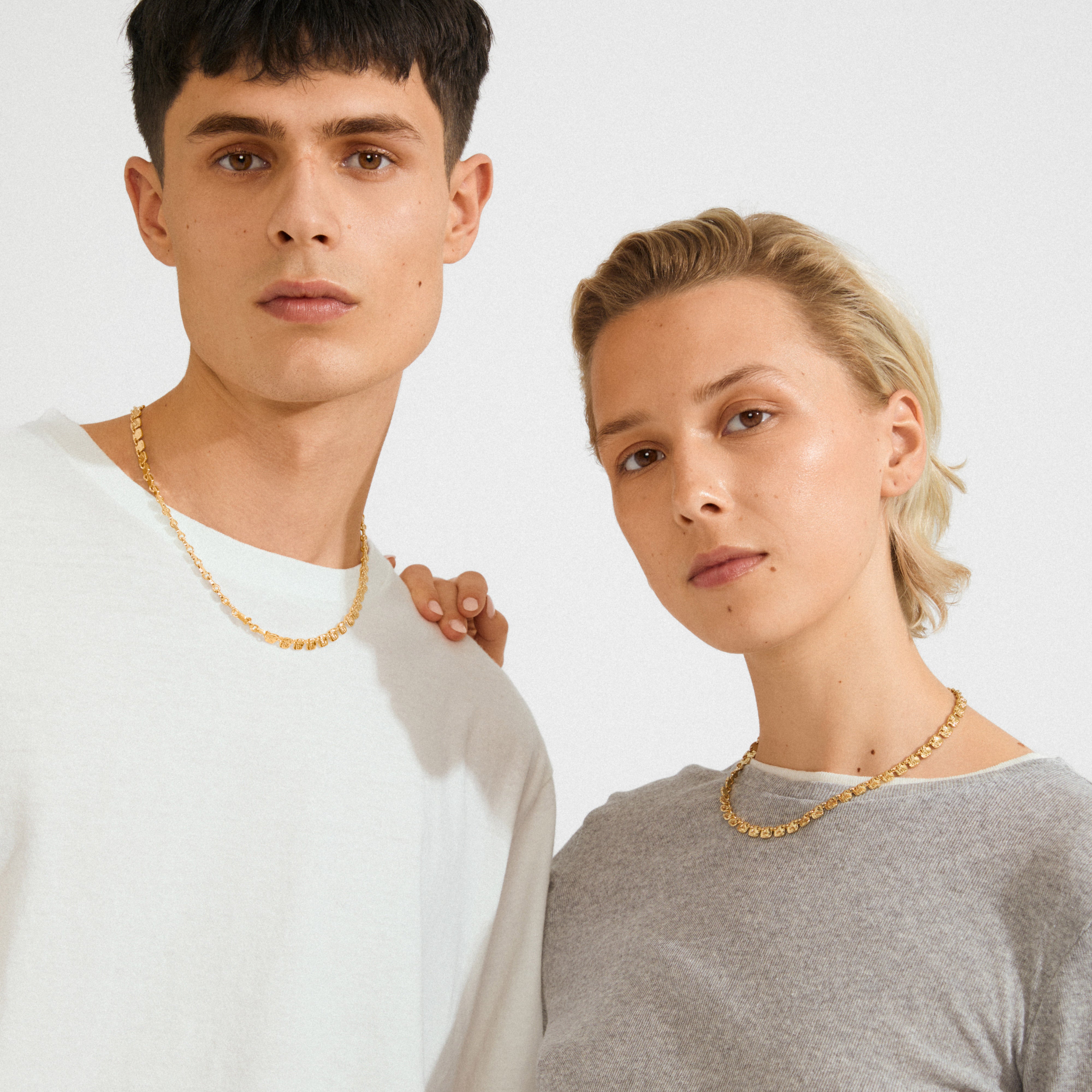 Two models showcasing Pilgrim's SENSE gold-plated necklace, featuring square coins with a raw texture, reflecting light for a stylish glow.