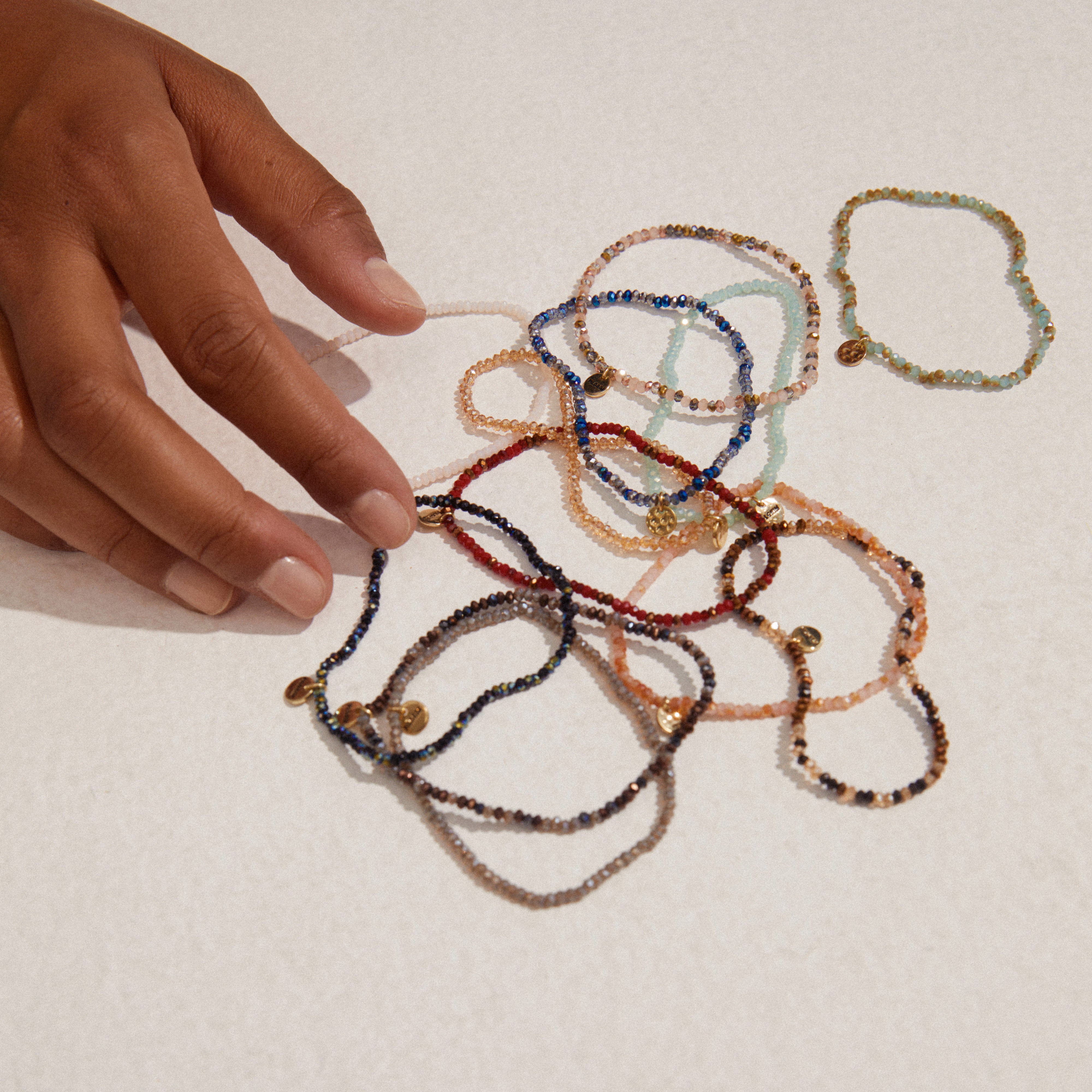 Hand holding the INDIE bracelet, featuring white, faceted glass beads with a gold-plated Pilgrim logo. Strung on durable elastic, fits most wrists.