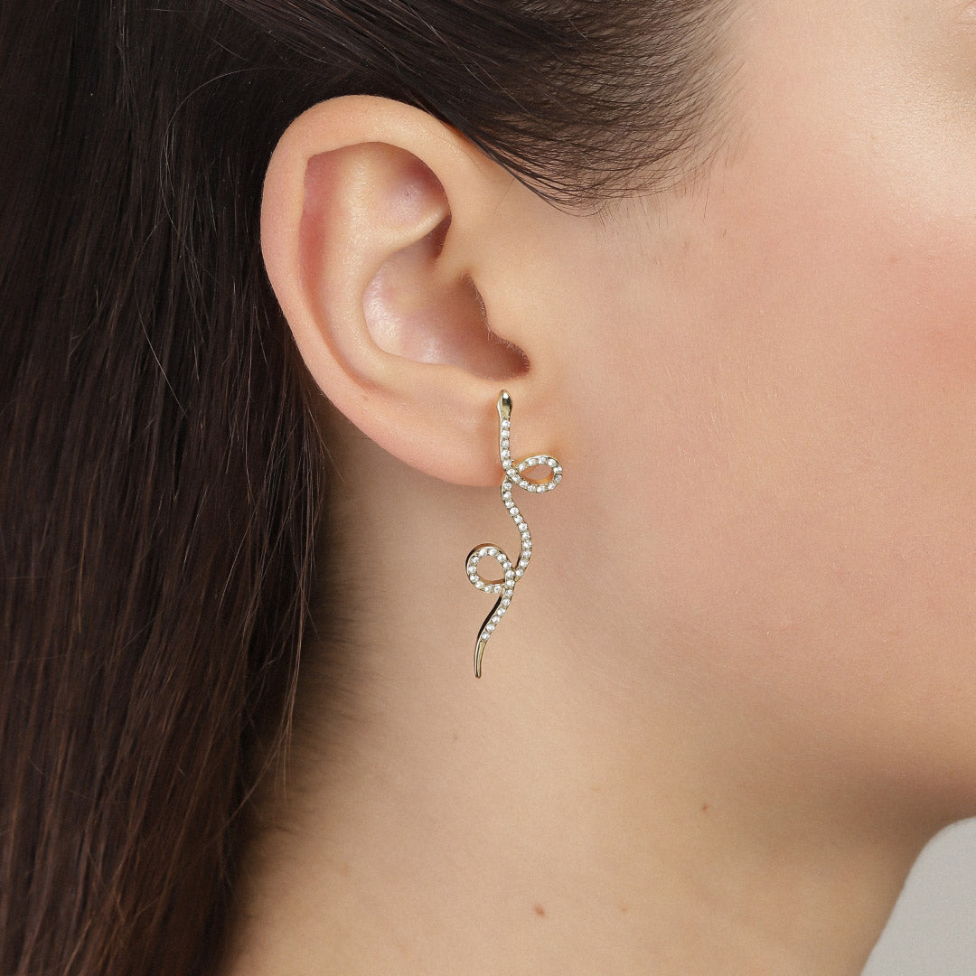 EBBA gold-plated earrings feature crystal-encrusted snakes elegantly adorning a woman's ear, embodying Pilgrim's blend of timeless and trendy Scandinavian jewelry design.
