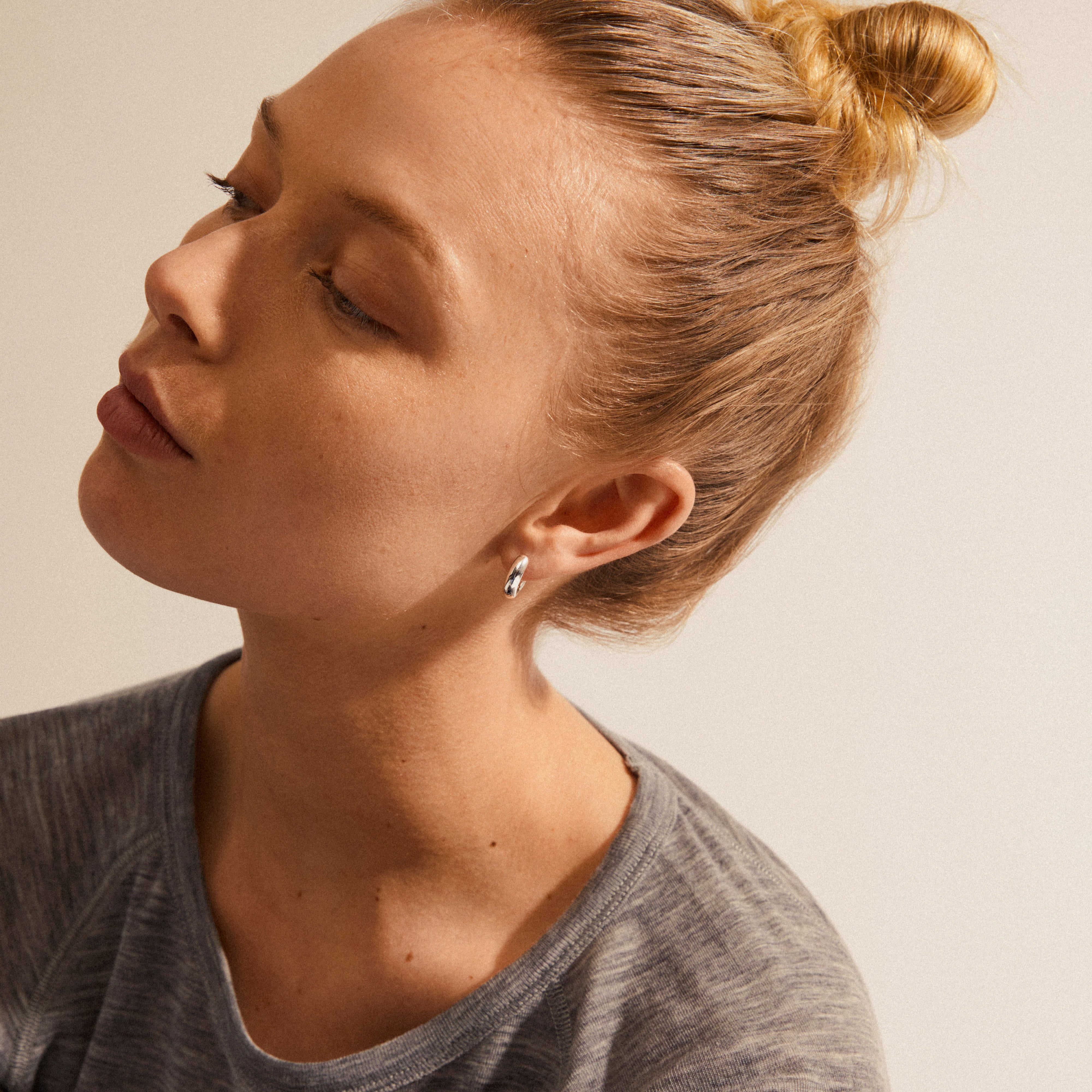 Woman wearing AICA chunky huggie hoop earrings, silver-plated, showcasing their minimalist design and shiny surface. Her eyes are closed, highlighting the earrings.