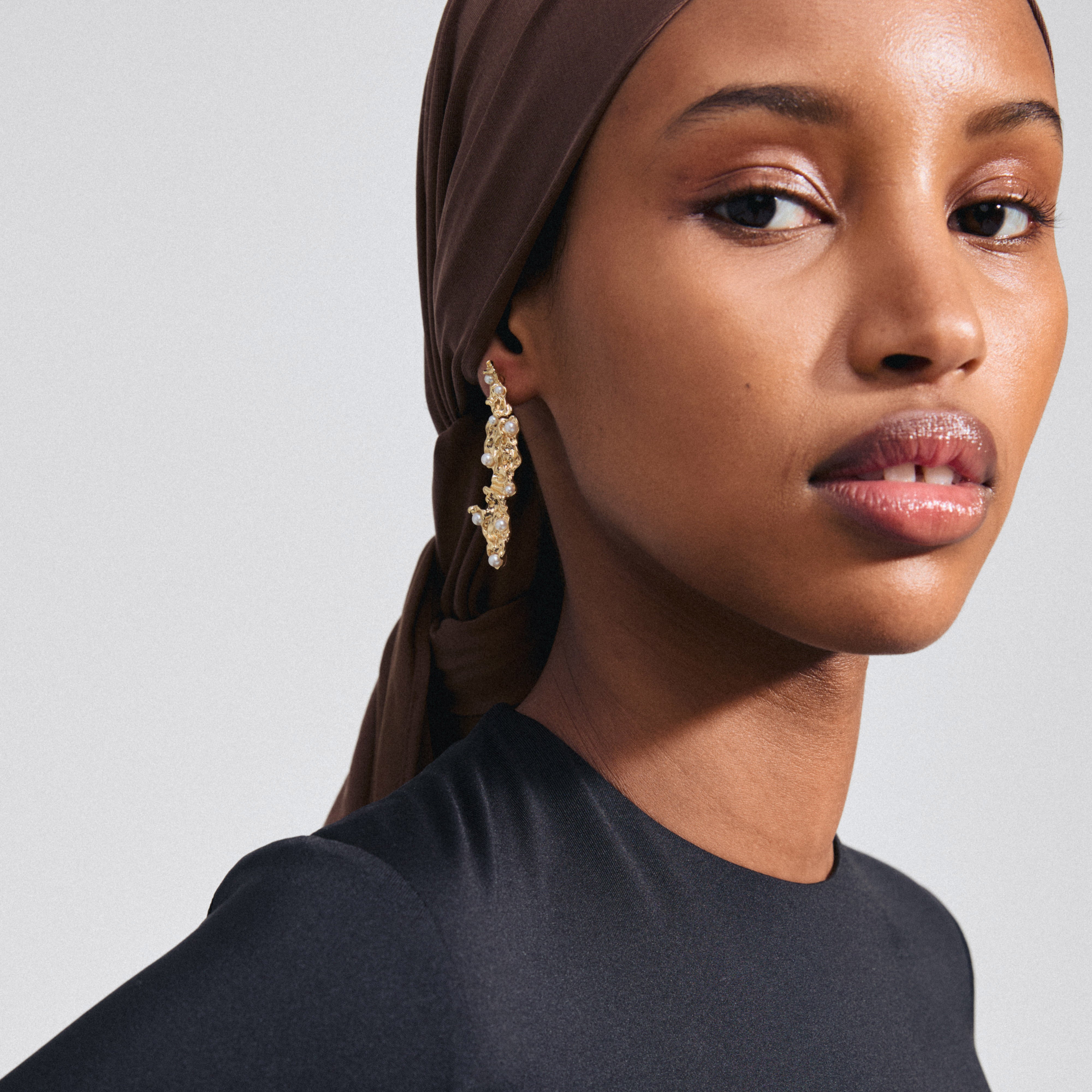 Close-up of JUDE earrings, gold-plated with organic design and pearlescent glass beads, worn by a woman with a headscarf, highlighting Pilgrim's unique style.
