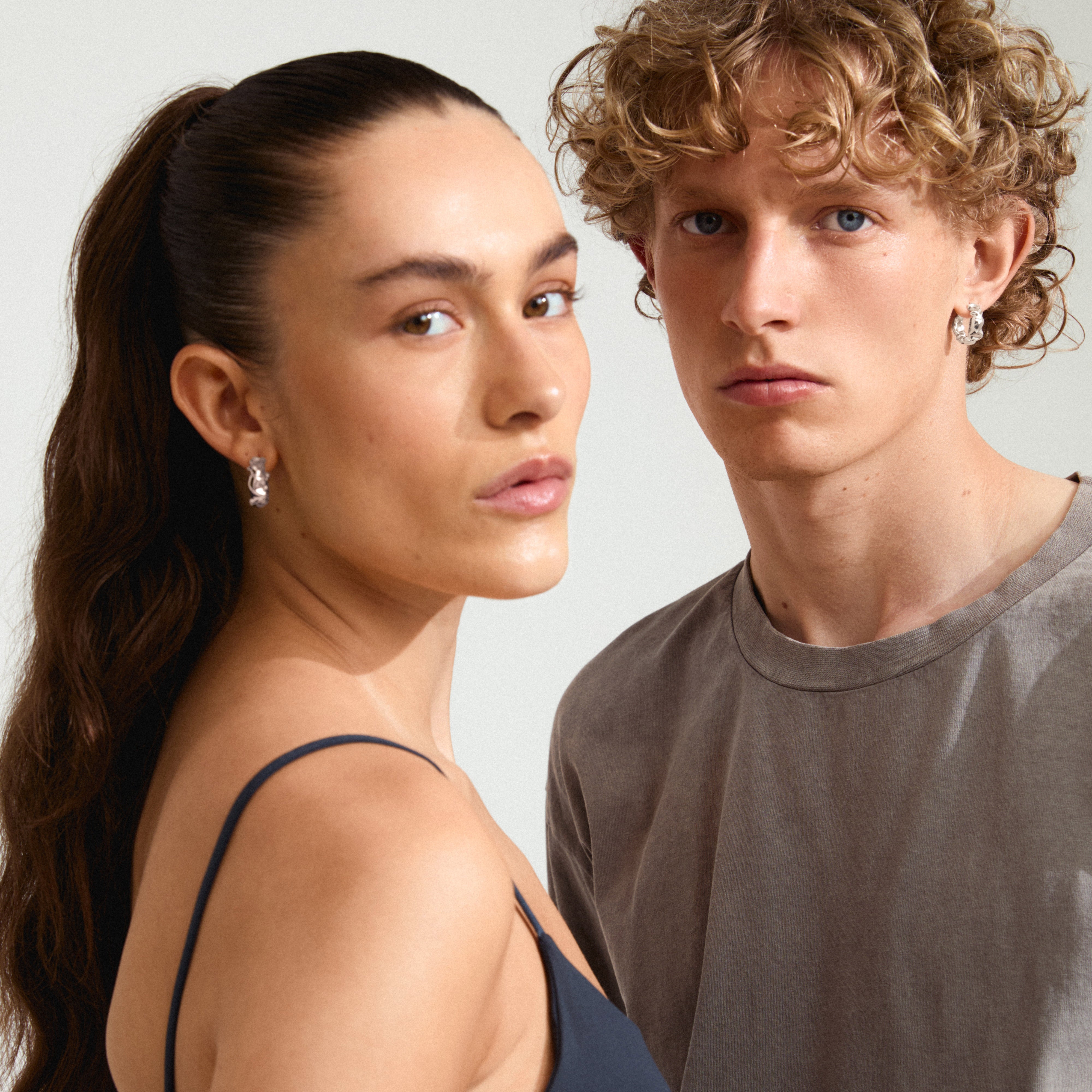 Two young adults model the ELLIOT silver-plated hoop earrings, showcasing their wavy design and modern elegance.