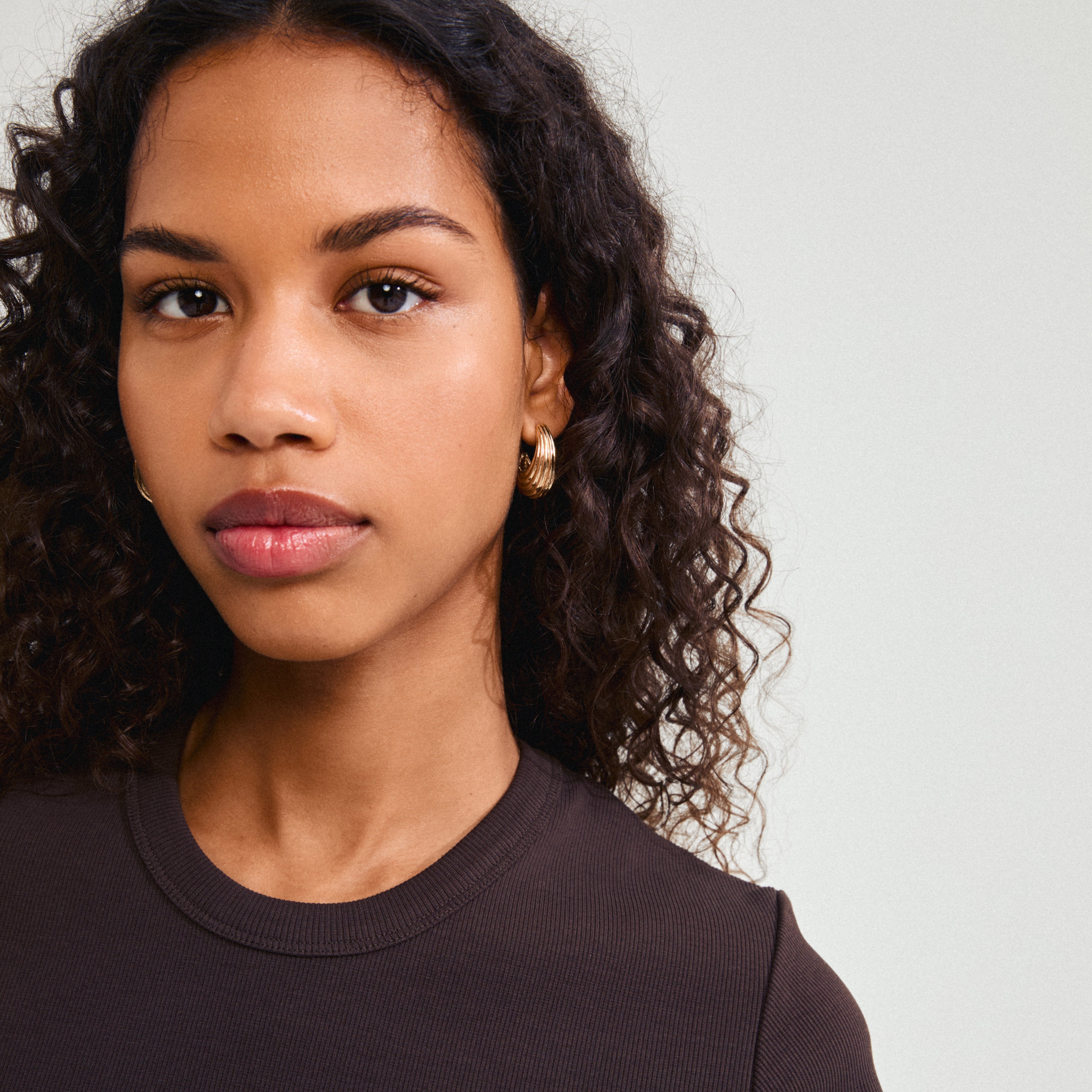 Woman with curly hair wearing ASLE gold-plated earrings, showcasing chunky, ribbed, half-hoop design by Pilgrim, inspired by ocean waves and sand dunes.