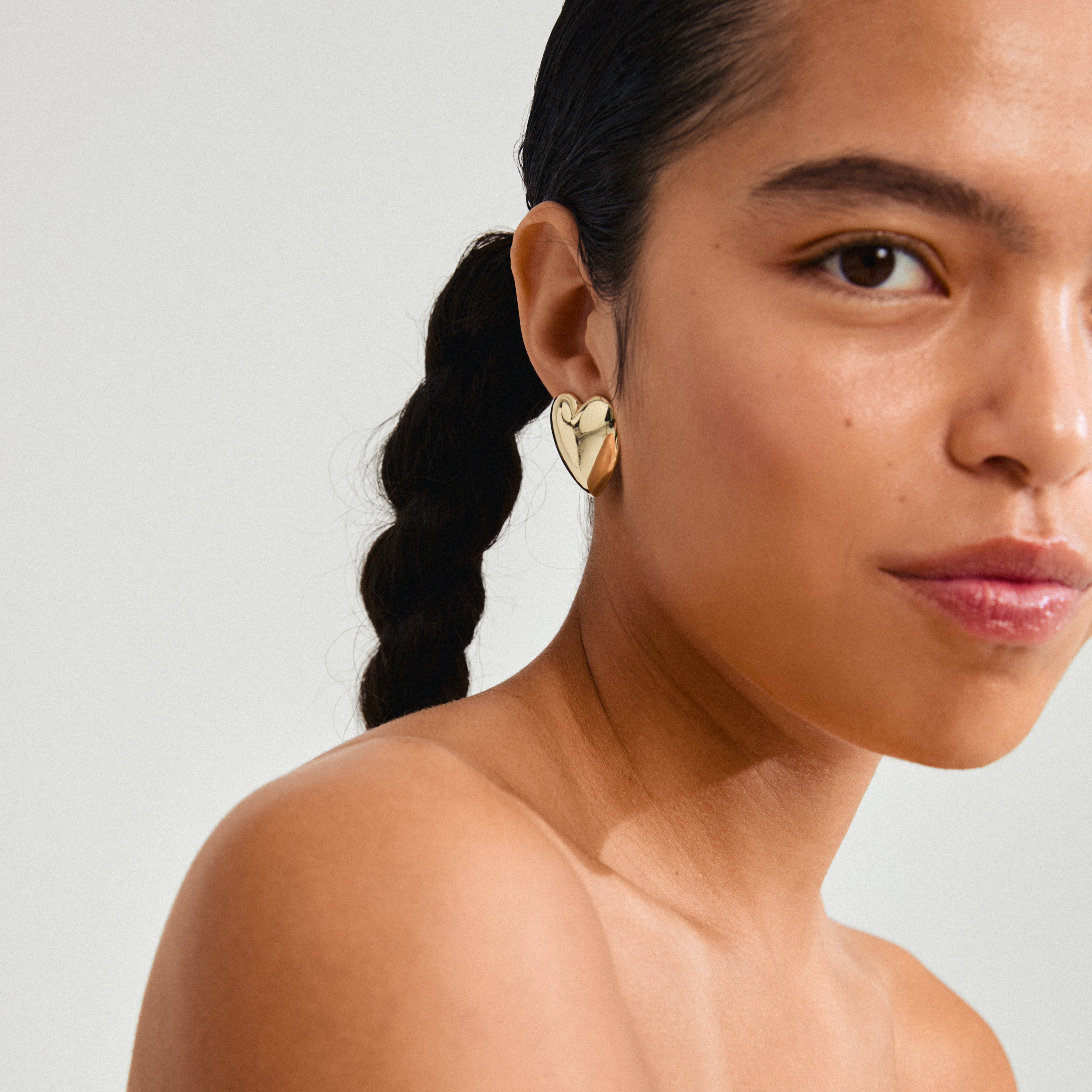 Close-up of a woman wearing Pilgrim's gold-plated ANNEMETTE earrings, showcasing chunky heart-shaped design with a shiny finish.