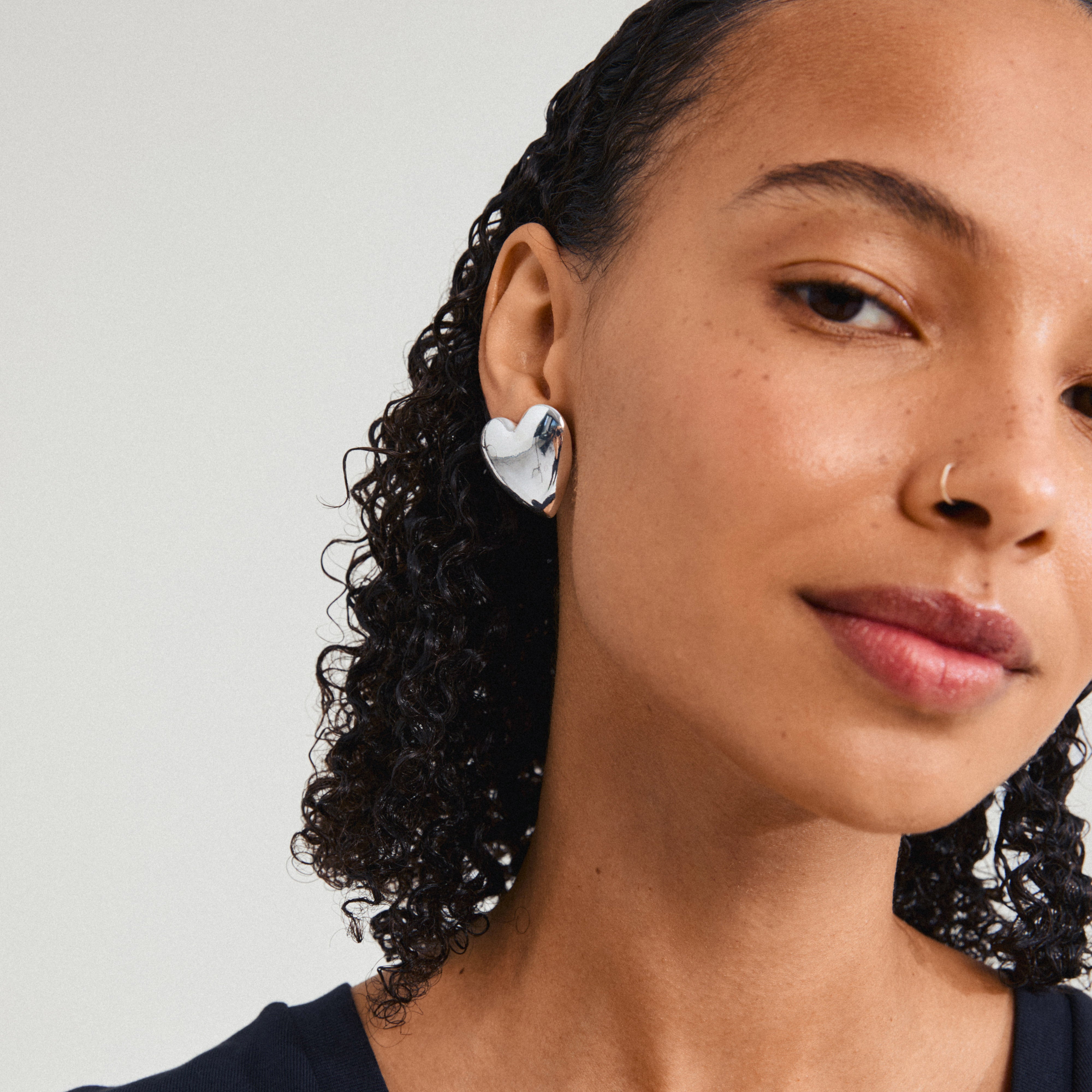 Close-up of a woman wearing silver-plated ANNEMETTE earrings, showcasing chunky heart-shaped design and shiny finish, by Pilgrim.