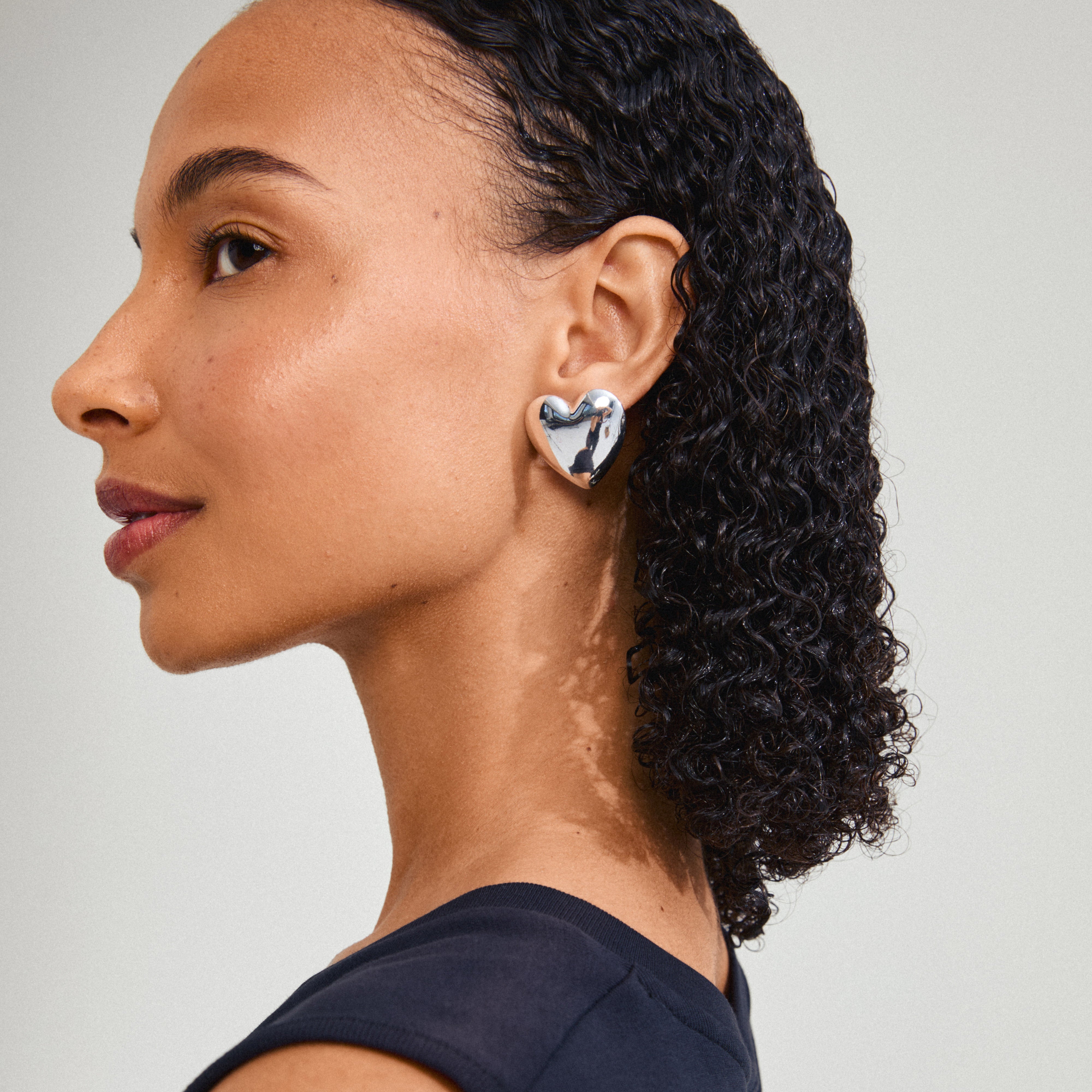 A woman with curly hair wears silver-plated ANNEMETTE earrings by Pilgrim, featuring chunky heart shapes with a shiny finish.