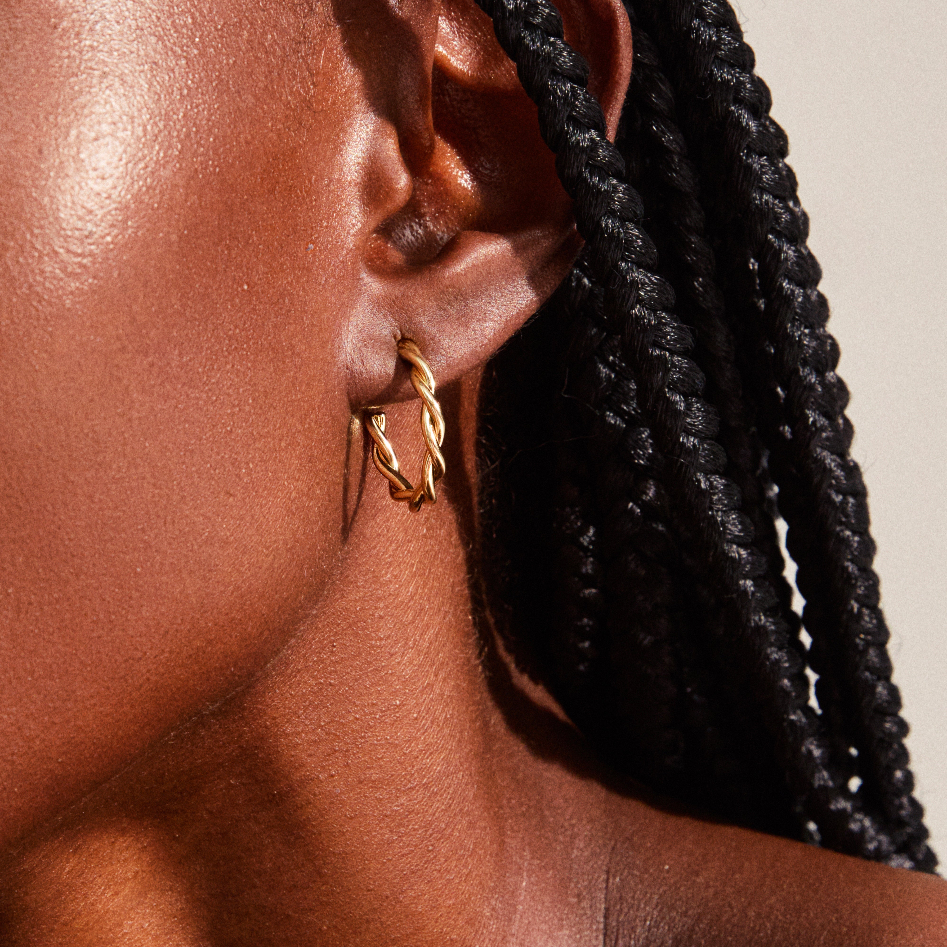 NAJA twisted hoop earrings in gold-plated design, shown on an ear with dark braided hair, highlighting the elegant, light-catching twisted details.
