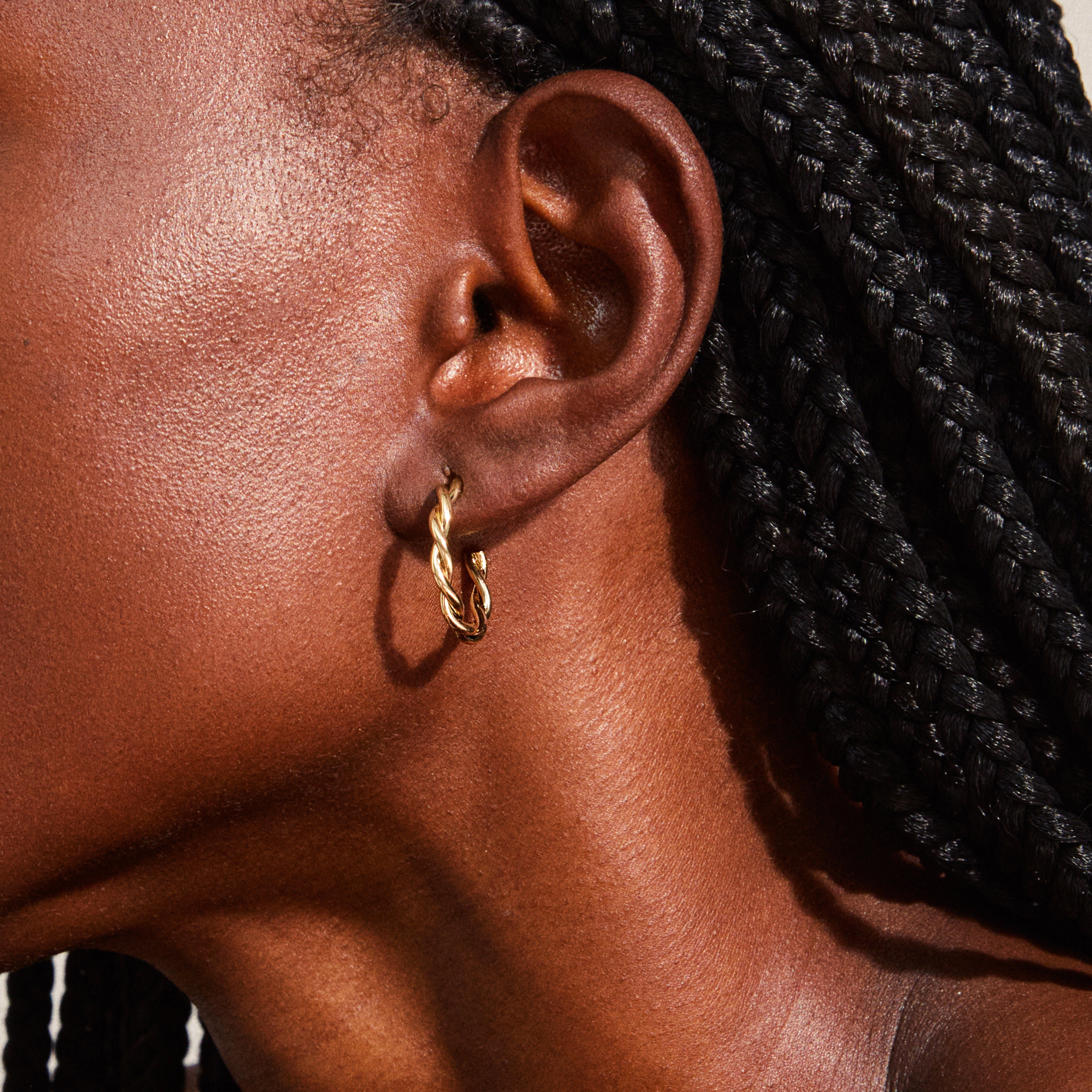 Close-up of NAJA twisted hoop earrings, gold-plated, adorning a person’s ear with neatly braided hair, showcasing elegant twisted-rope design.