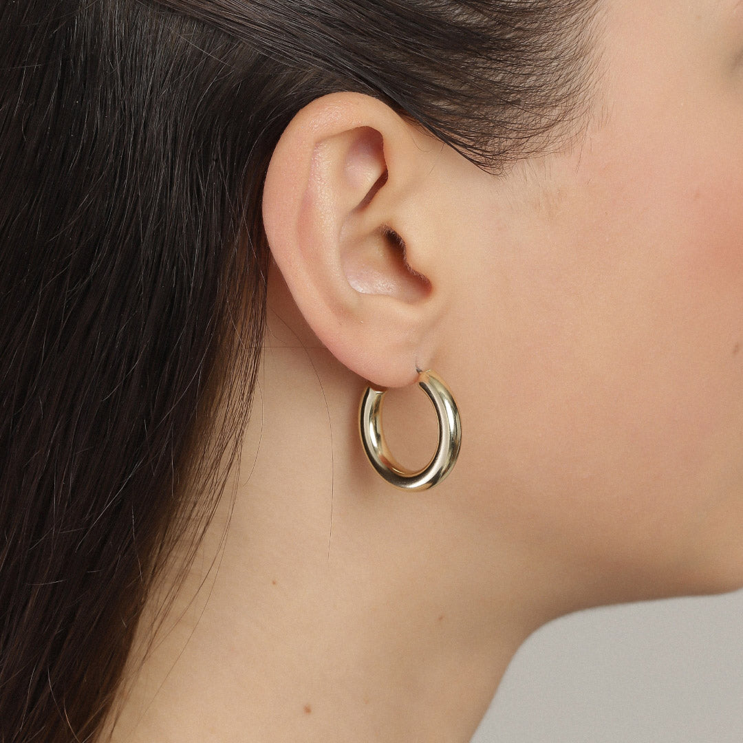 MADDIE chunky gold-plated hoop earrings adorn a woman's ear, showcasing their shiny, bold design perfect for elevating both casual and elegant styles.