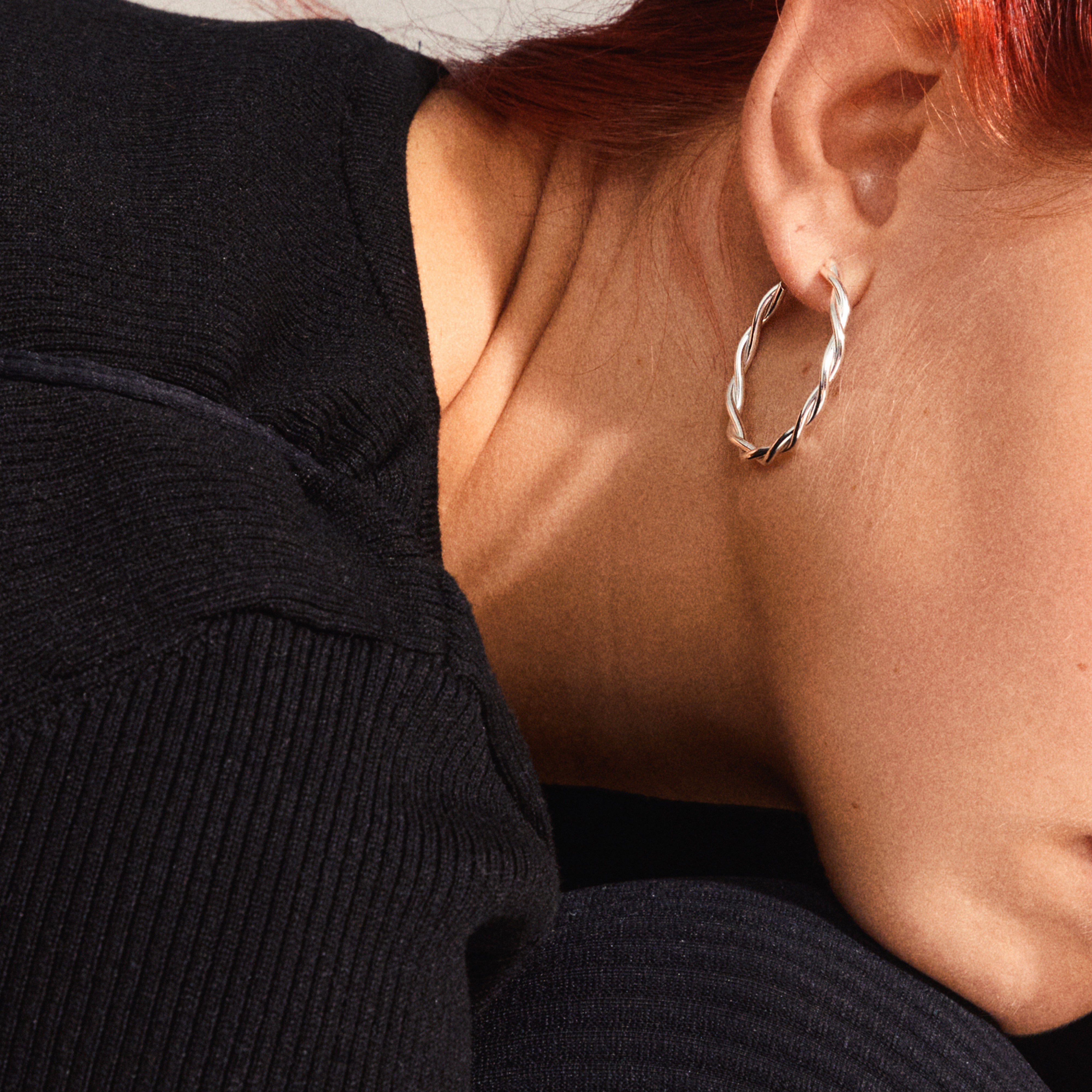 Close-up of a person wearing NAJA large hoop earrings, silver-plated with twisted-rope design, showcasing chic reflections, enhancing the black ribbed top.