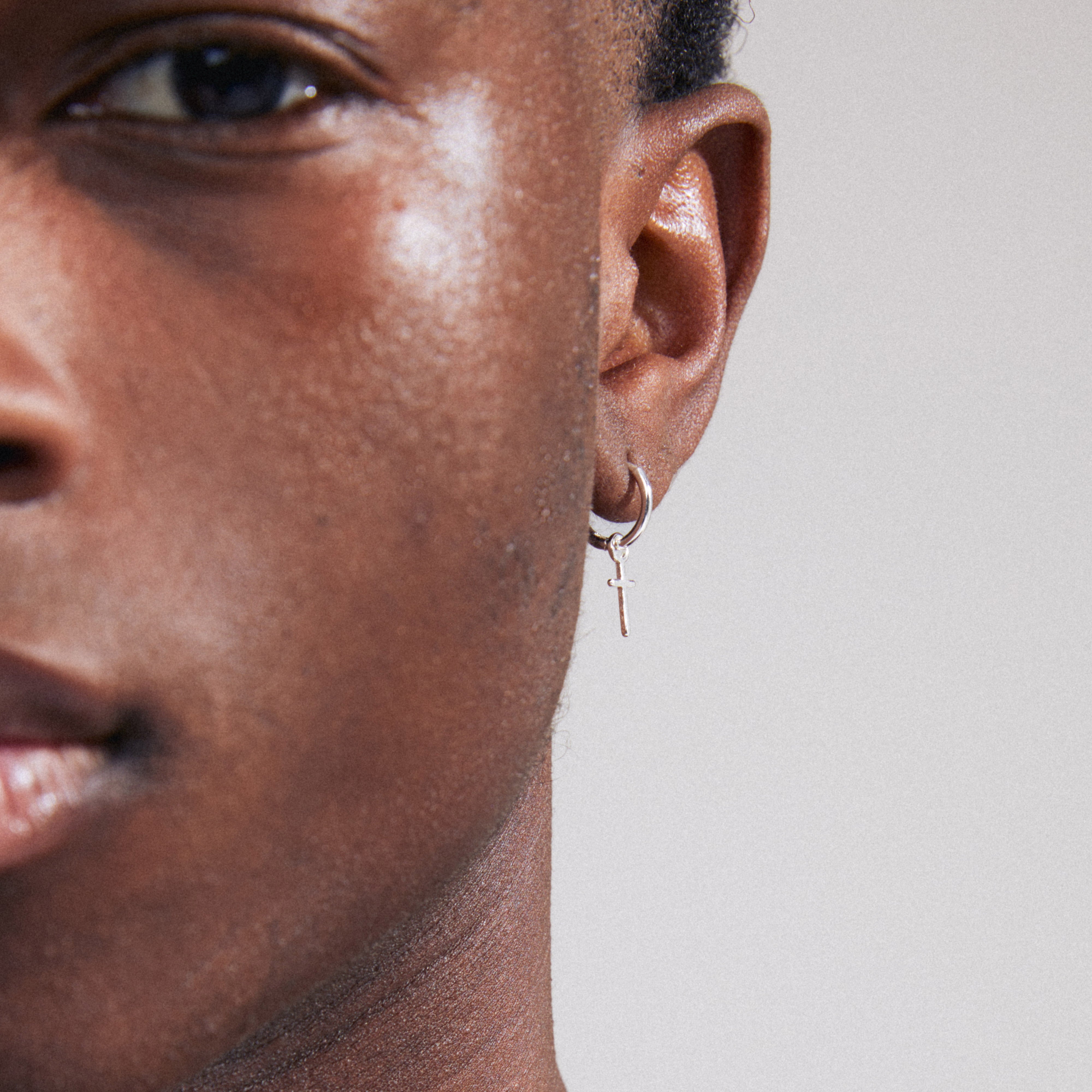 XENAIA single earring silver-plated, featuring a small hoop with a cross pendant, shown on a person's ear. Timeless design with a shiny finish.