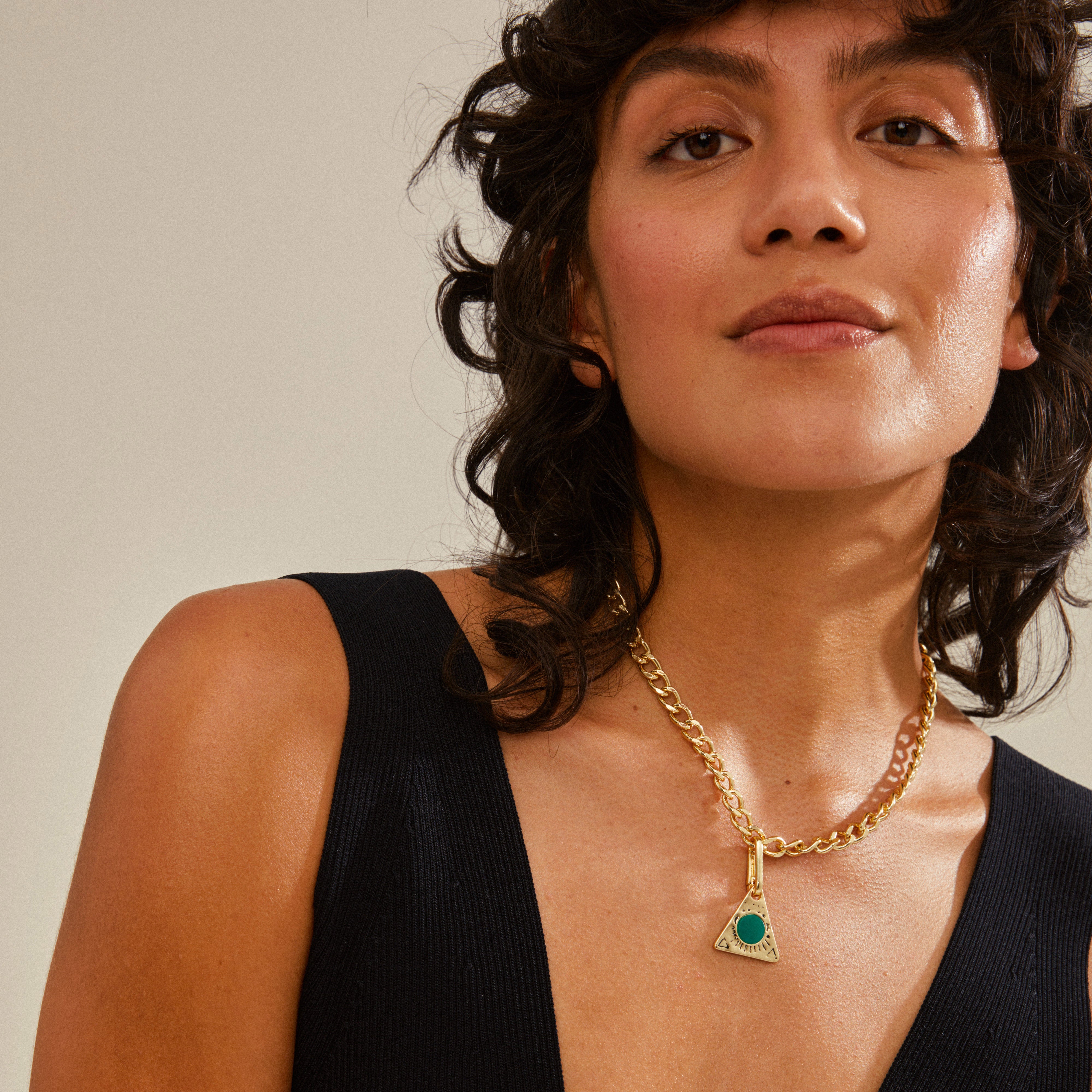 A woman wears Pilgrim's CHARM triangle pendant, green/gold-plated, styled on a gold chain, showcasing its elegance and personalization potential.