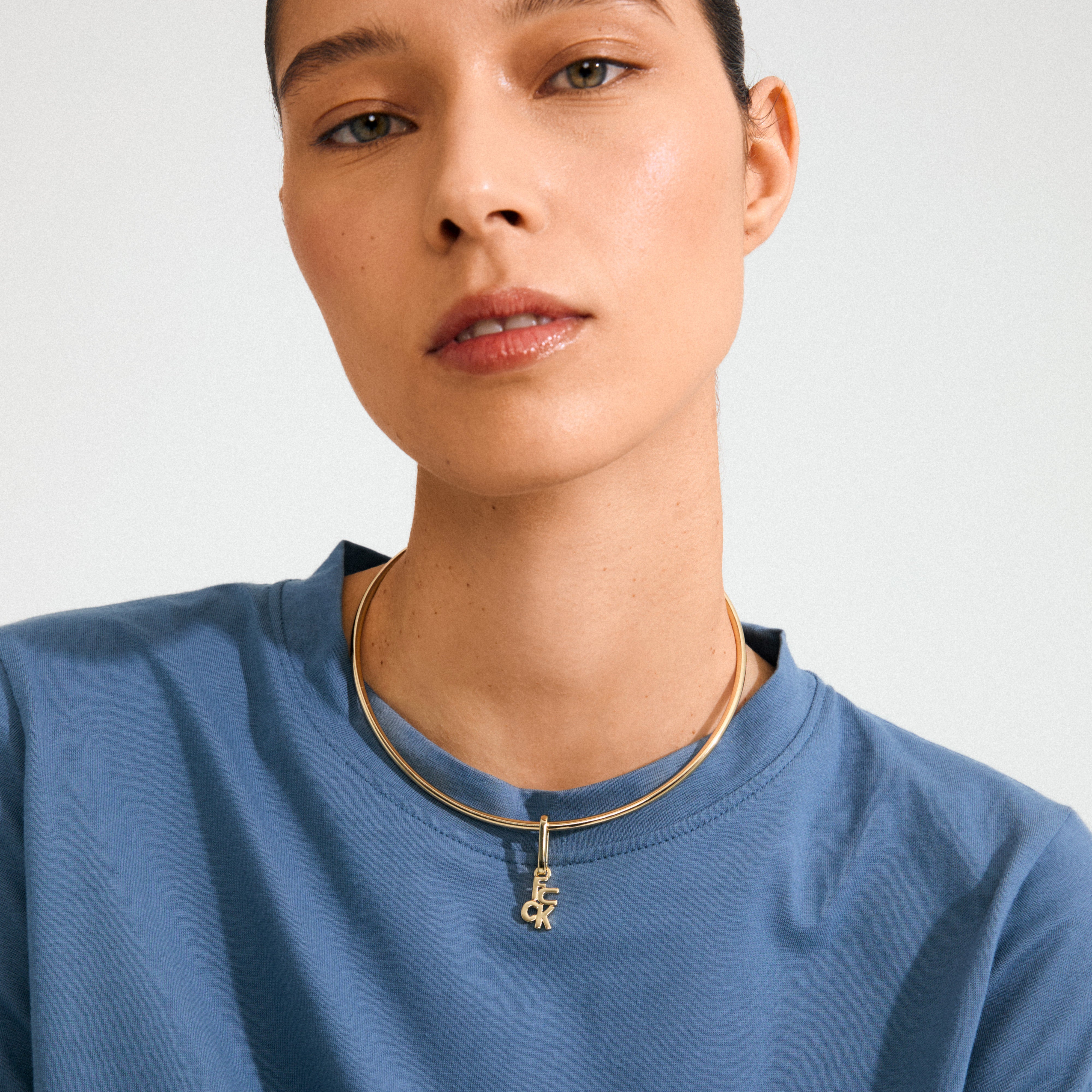 Close-up of a person's neck wearing a sky-blue T-shirt and the CHARM f... pendant, gold-plated, on a rigid gold choker, showcasing Pilgrim's edgy style.