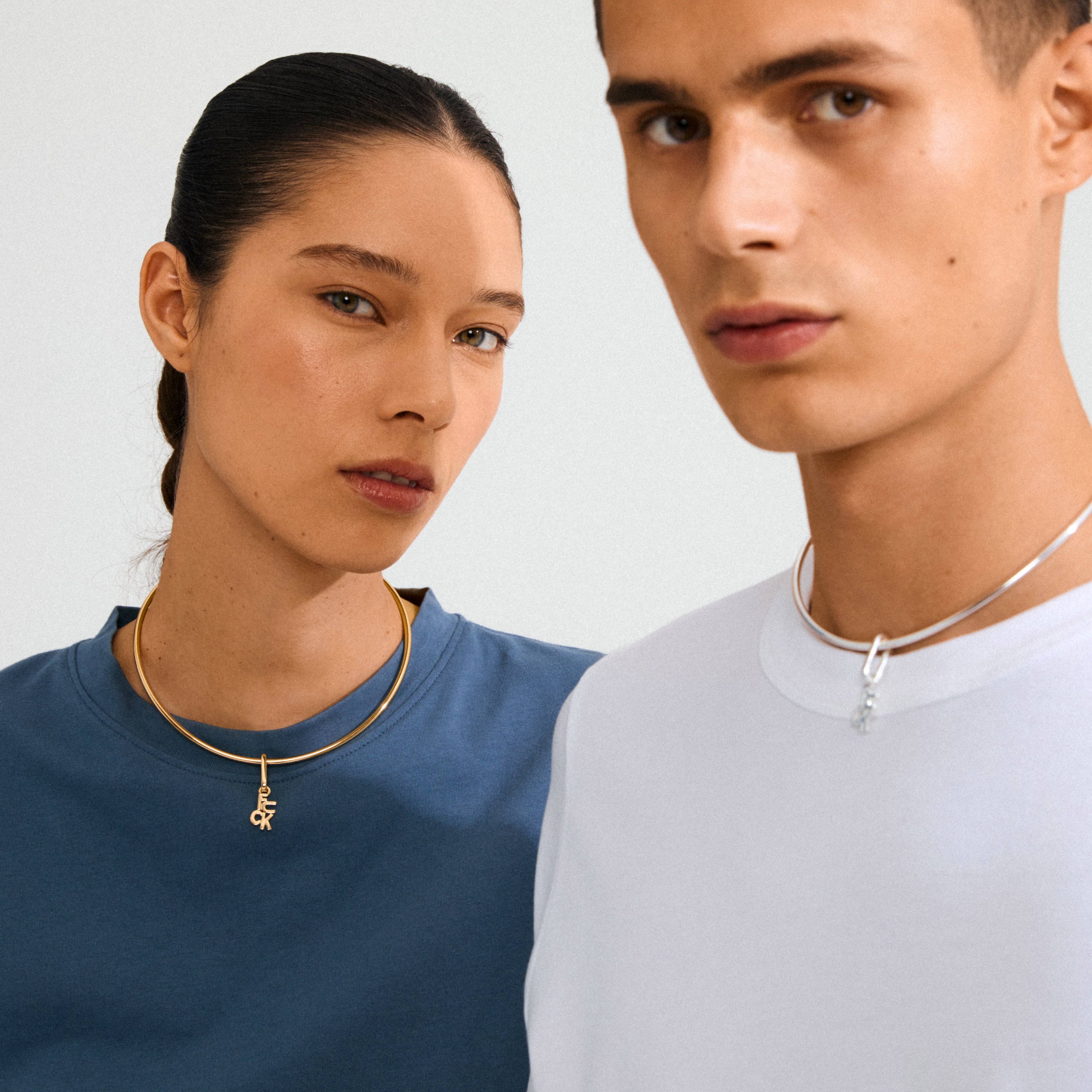Two young adults wearing simple crewnecks showcase Pilgrim's CHARM f... gold-plated pendant, emphasizing minimalistic, bold personal style with a recycled material focus.
