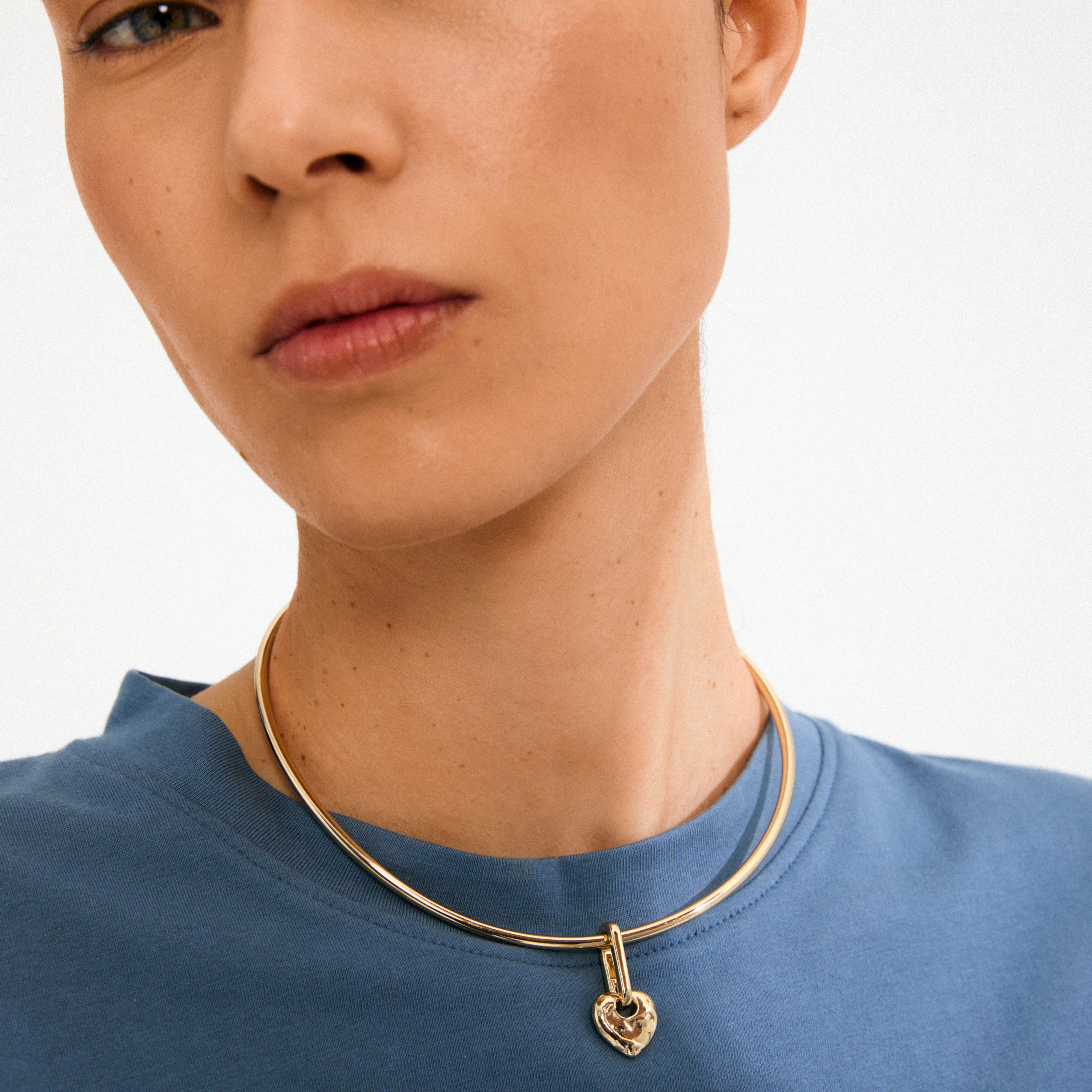 Person wearing a simple blue top with Pilgrim's CHARM rough heart pendant, a unique gold-plated choker-style necklace featuring a rustic heart-shaped padlock.