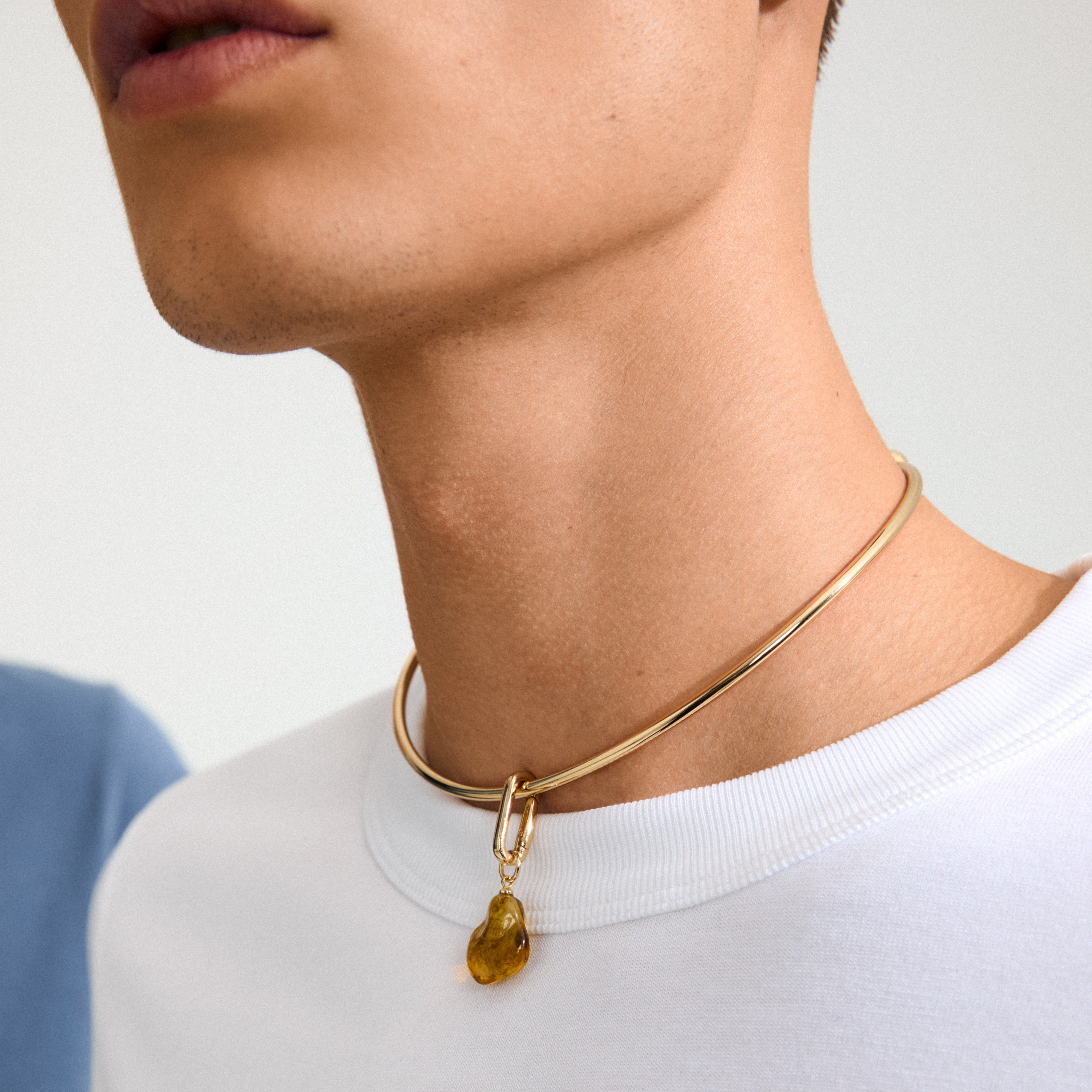 Close-up of Pilgrim's CHARM mold pendant: a gold-plated choker with an organic-shaped amber pendant, worn over a plain white T-shirt.