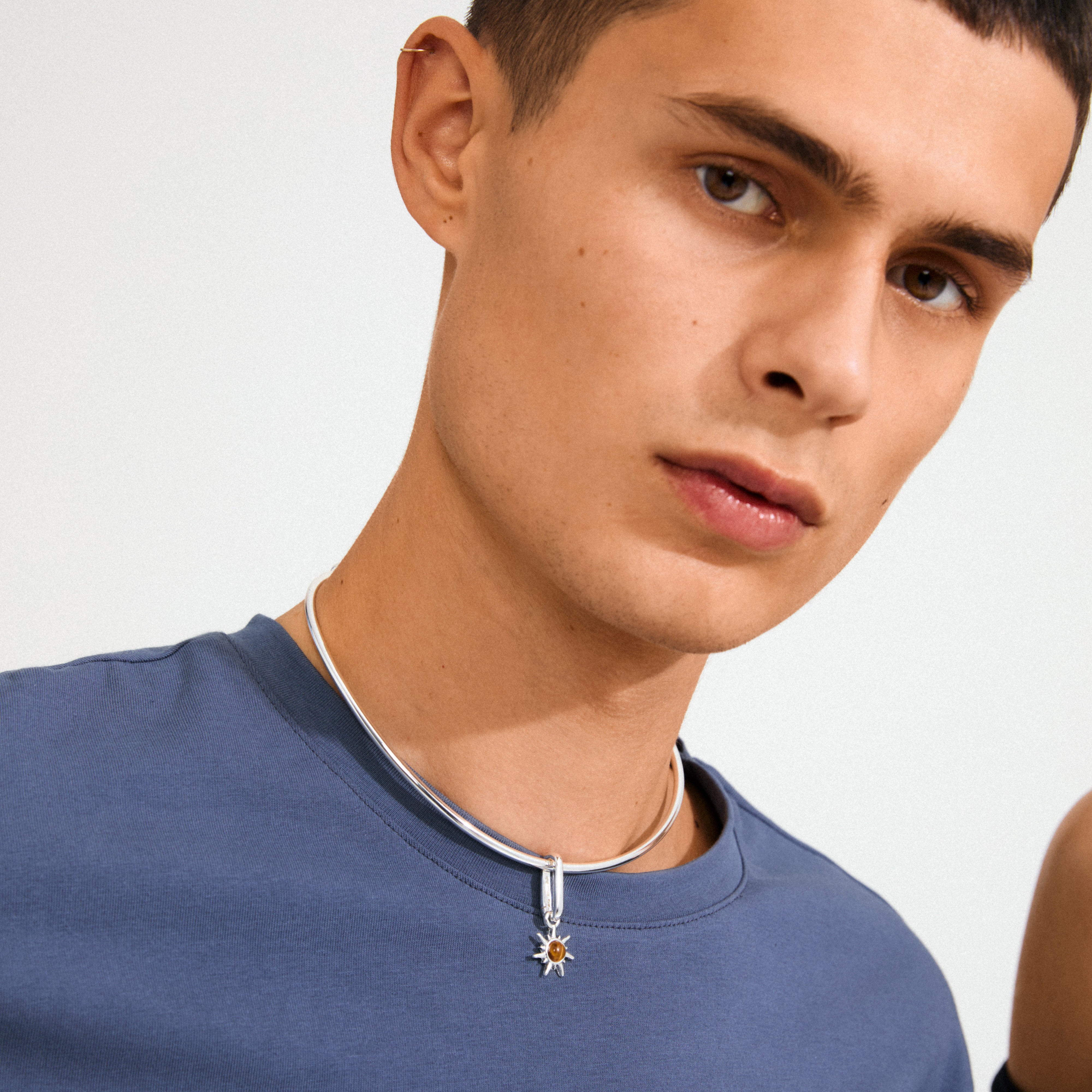 Young man wearing Pilgrim's CHARM bright pendant, silver-plated, with a Tiger Eye stone, highlighting its stylish sun-star design against a simple blue crew-neck T-shirt.