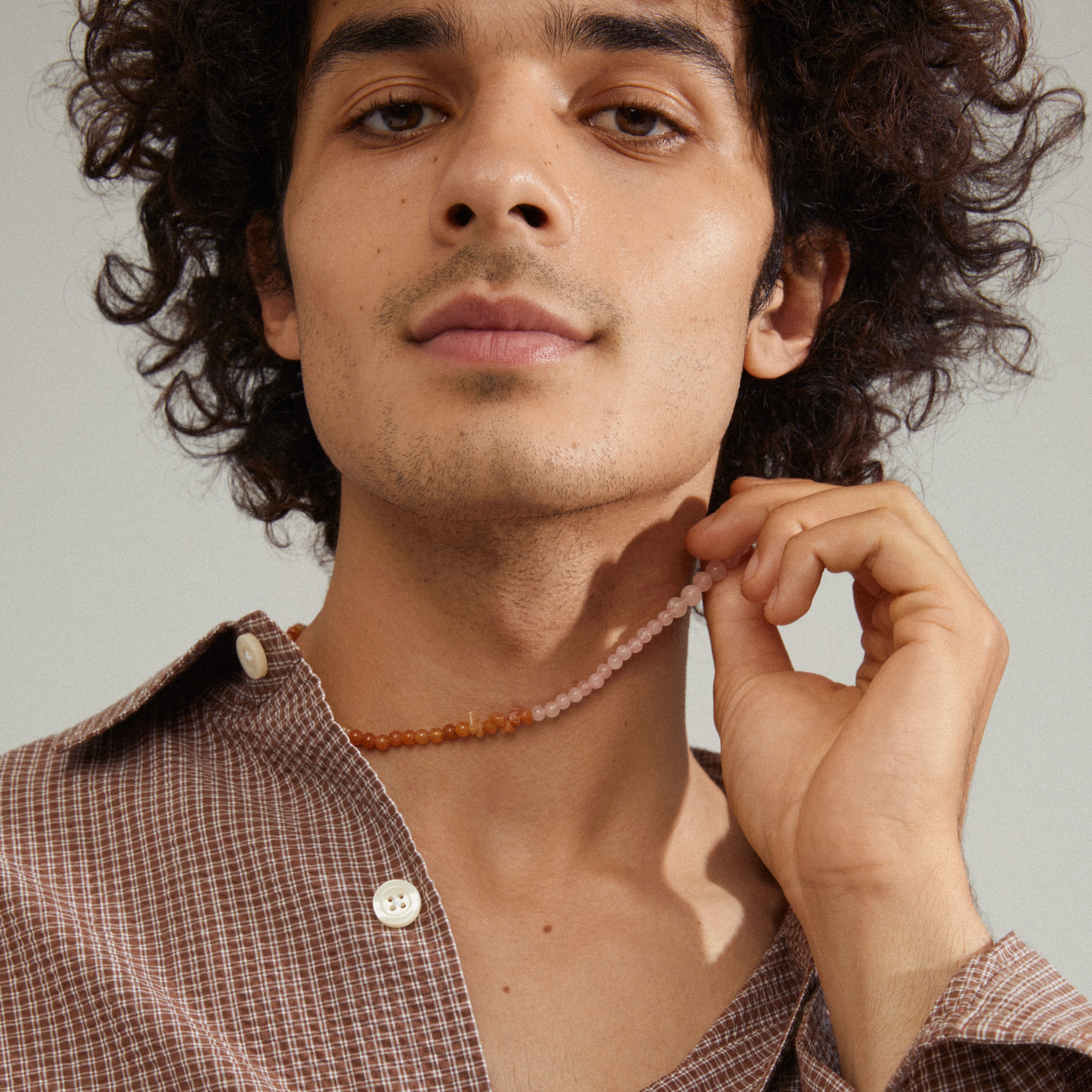 Man with curly hair holding the SOULMATES necklace, featuring rose quartz and red aventurine beads, representing love and strength.