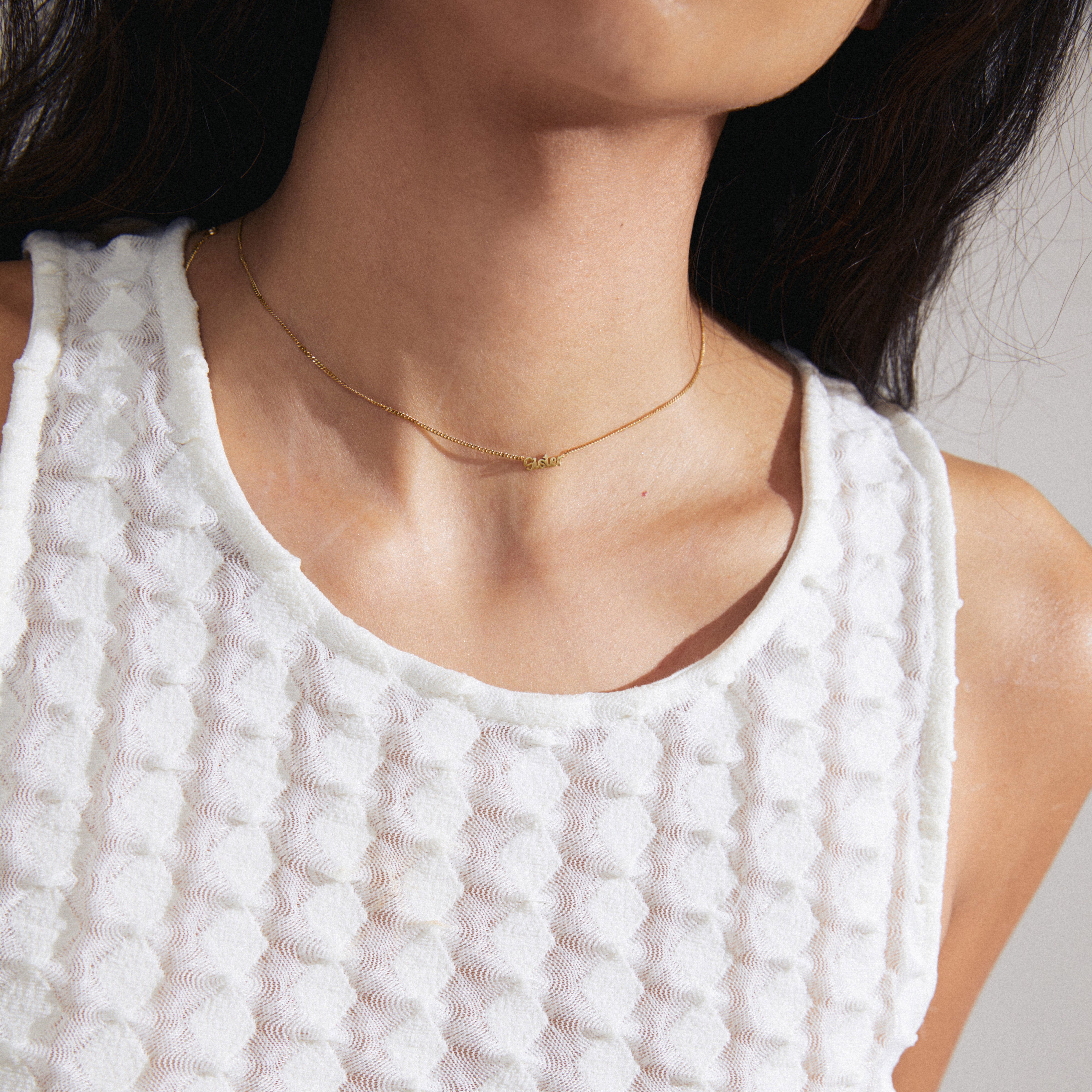 Gold-plated SISTER necklace worn by a woman in a white shirt, featuring a sleek pendant in cursive script, highlighting personal style and Scandinavian craftsmanship.