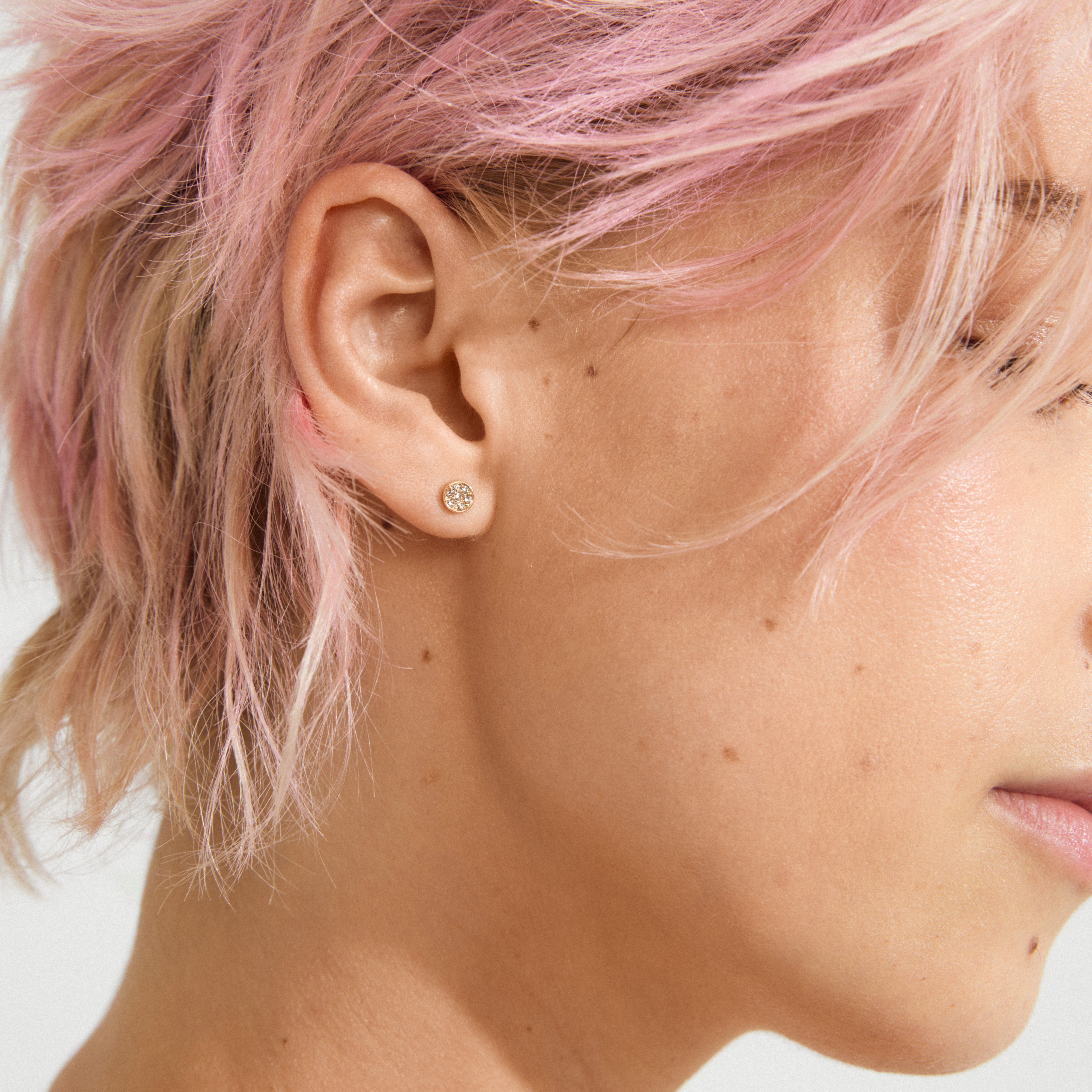 Side view of a person with pink hair wearing EMMA crystal earrings, gold-plated studs with sparkling stones, showcasing Pilgrim's blend of timeless and trendy Danish design.