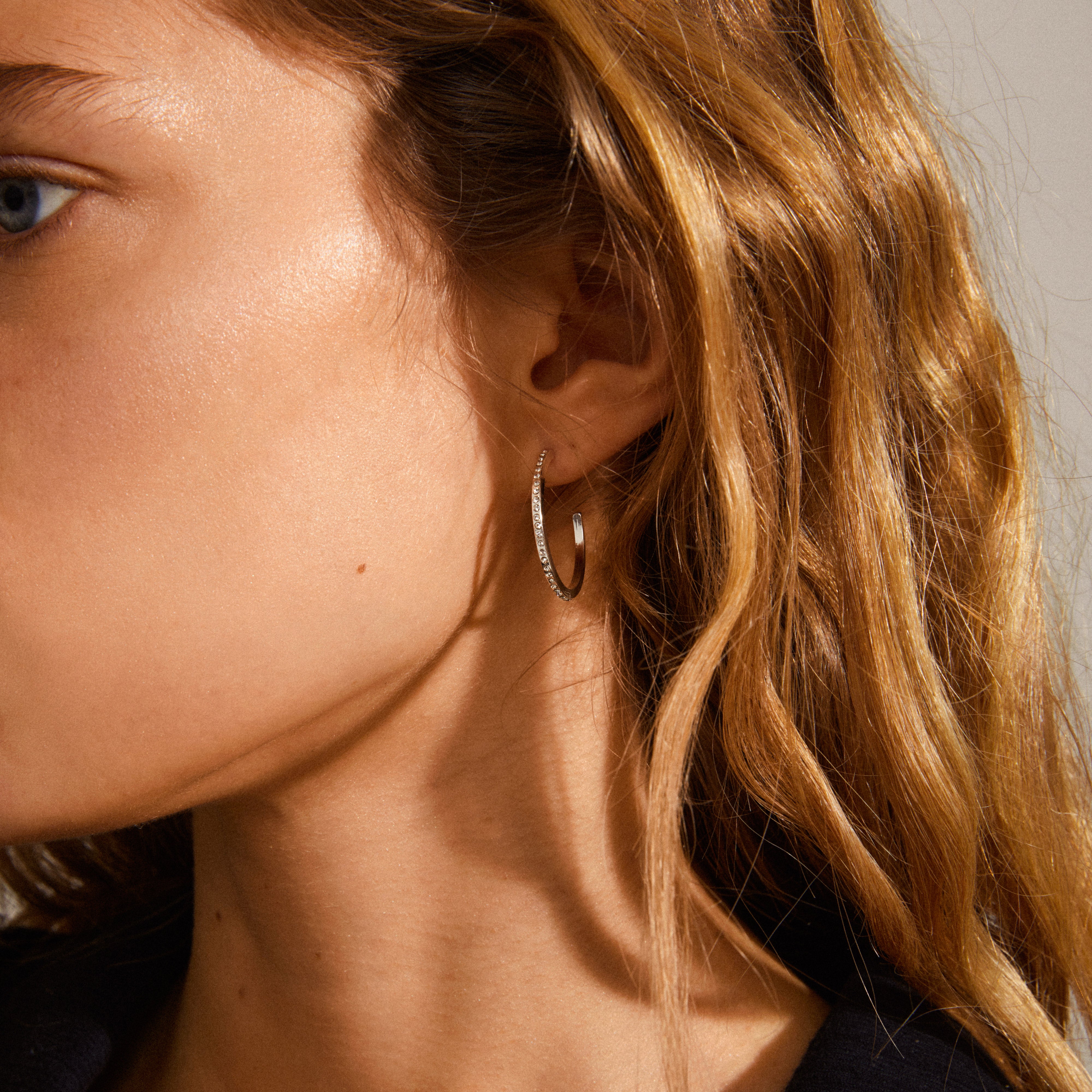 ROBERTA large crystal semi-hoops, silver-plated, elegantly adorn a woman's ear, showcasing their classic Nordic design with clear crystals for a chic, timeless look.