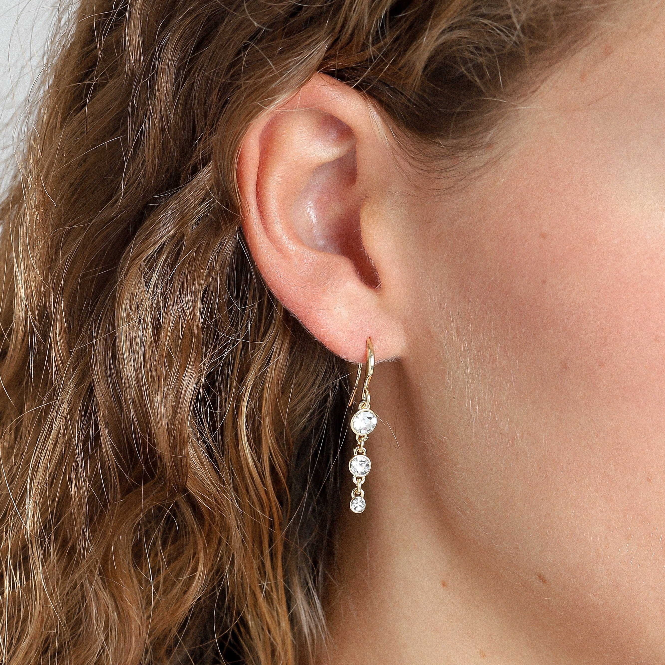 Close-up of a woman's ear wearing LUCIA gold-plated earrings with three sparkling crystals, showcasing a blend of timeless elegance and modern style.