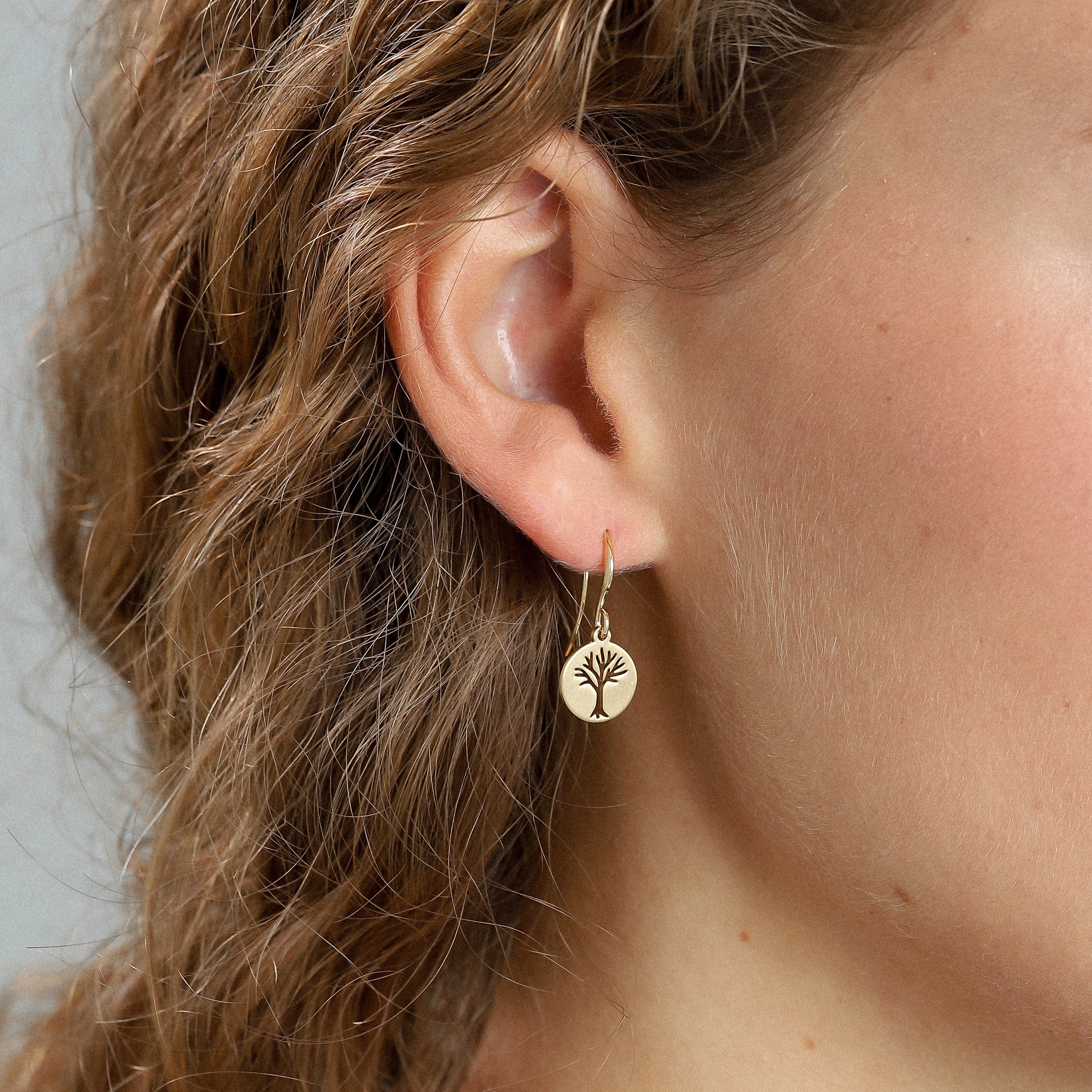 Gold-plated ELIN coin earrings featuring a minimalist tree design, showcased in a close-up of a woman's ear, highlighting their modern, trendy style.