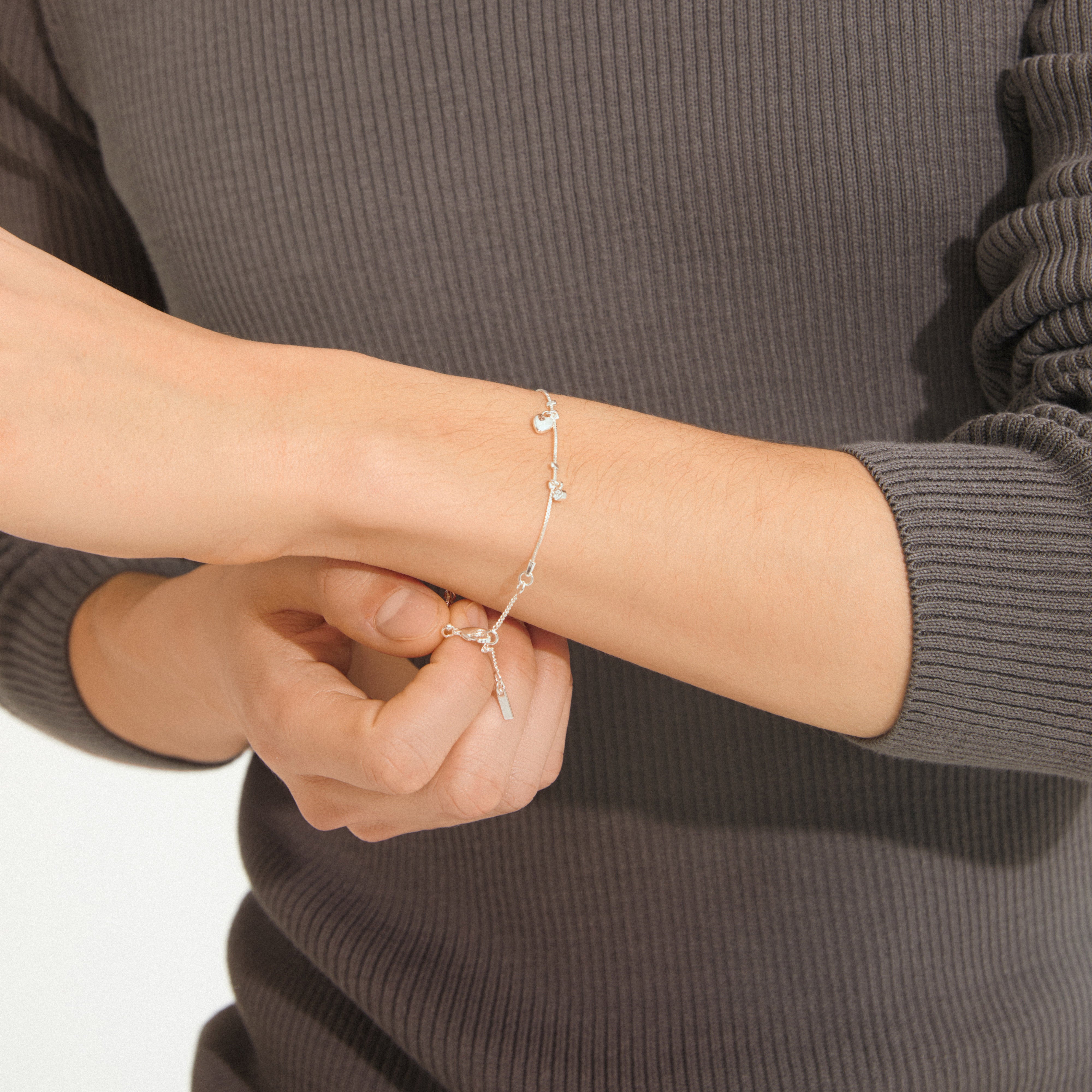 Hands fastening the ANET crystal bracelet silver-plated, featuring symbolic pendants for faith, hope, love, and Preciosa crystals, complementing a charcoal-gray ribbed top.