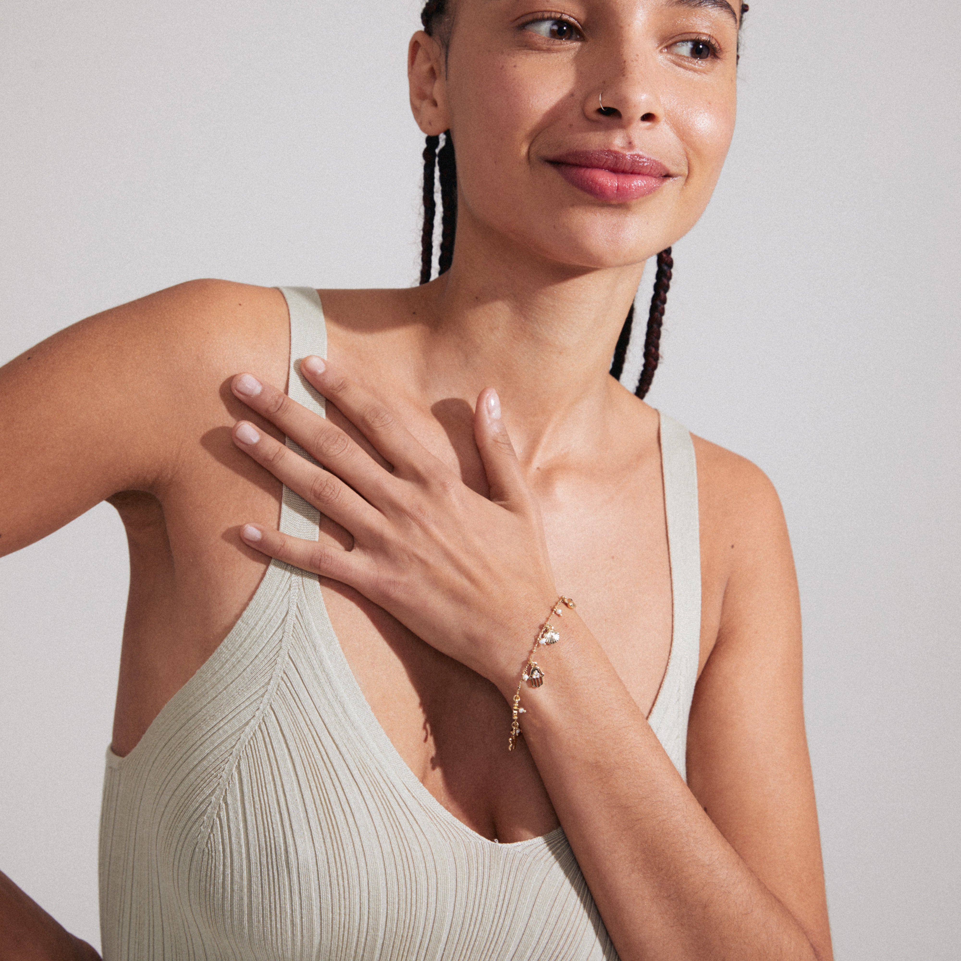 Woman with braids wearing the PRUCENCE gold-plated bracelet by Pilgrim, featuring charms and crystals, symbolizing love and dreams. Hand rests near chest, showcasing the bracelet.