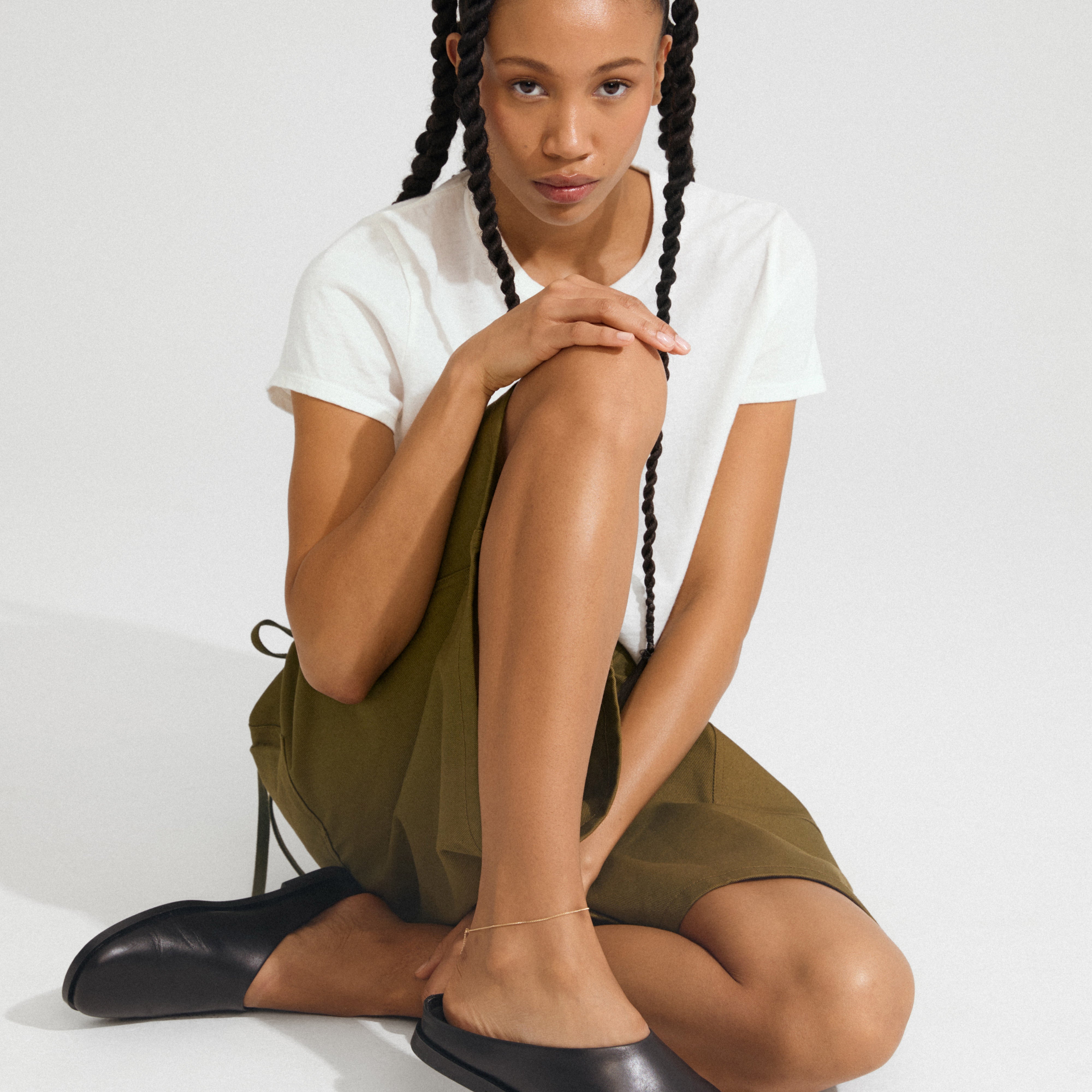 Young woman wearing the PALLAS anklet gold-plated on her right leg, paired with black mules and an olive-green skirt, sitting gracefully against a white backdrop.