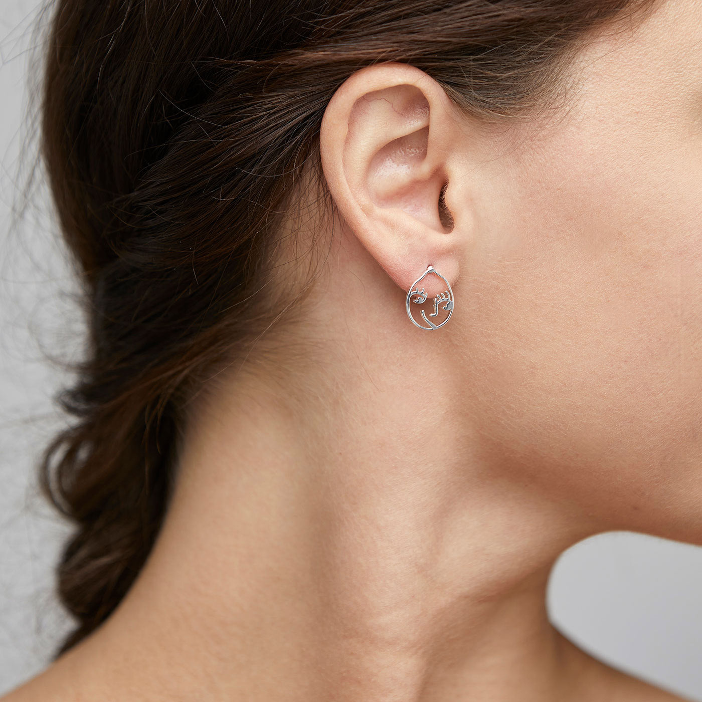 DEBRA earrings silver-plated on a woman's ear, featuring a smiling face motif in an irregular oval, showcasing a blend of artistry and unique style.