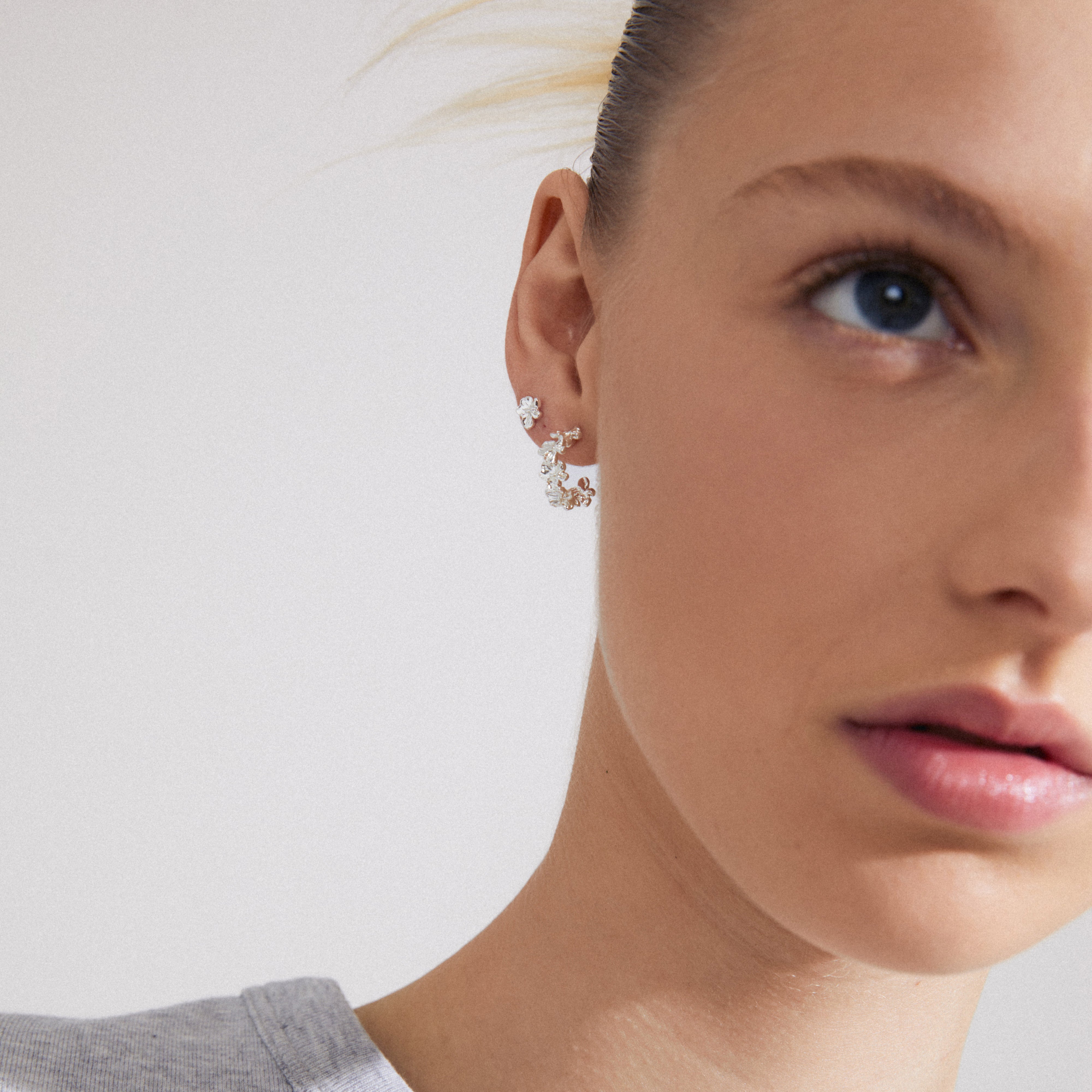 Close-up of a woman's ear wearing ZHURI earrings 2-in-1 set, featuring silver-plated floral half hoops and tiny floral studs from Pilgrim.