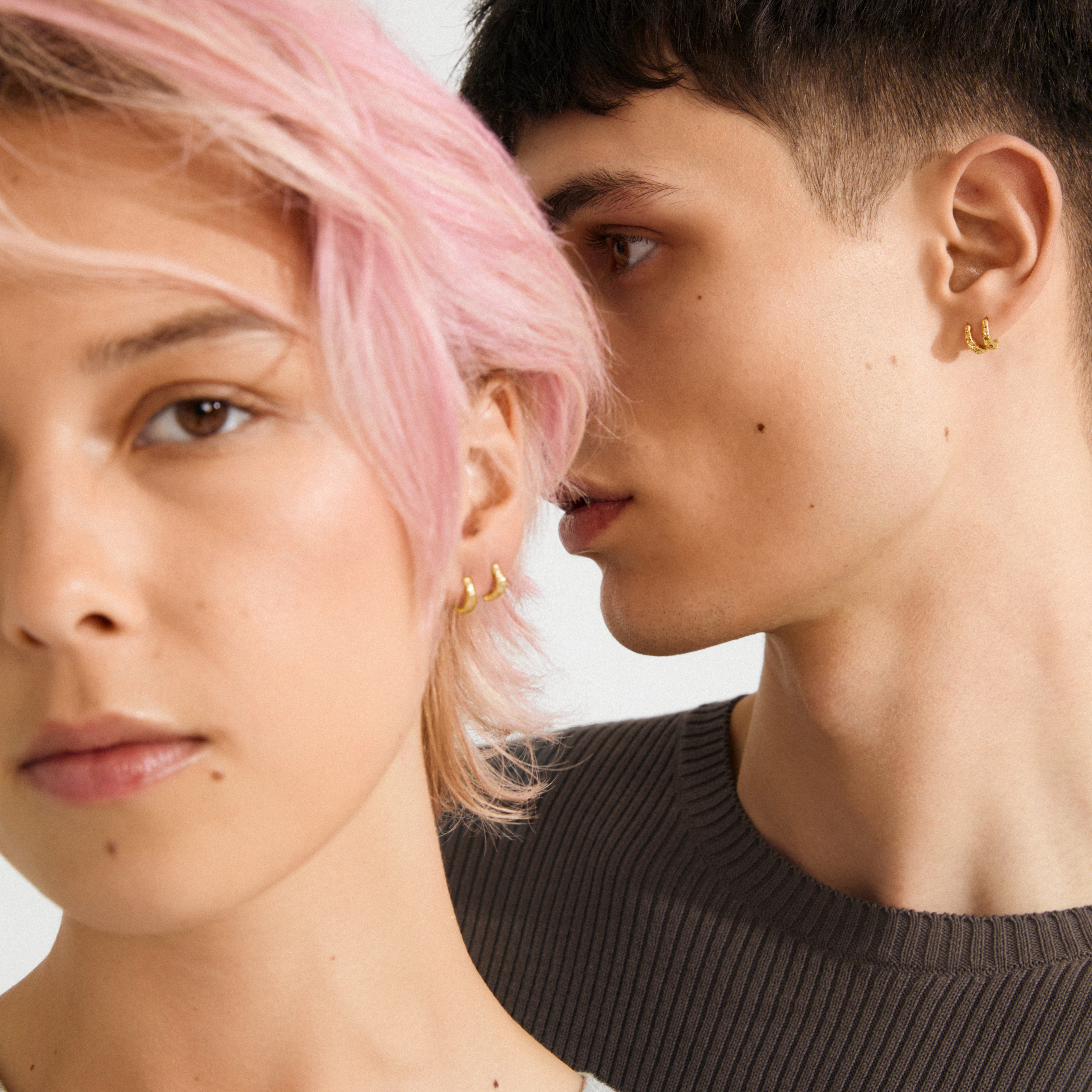 Two young adults showcase EDDY organic-shaped mini huggie hoops gold-plated, emphasizing their handcrafted texture and nature-inspired design, perfect for a curated ear look.