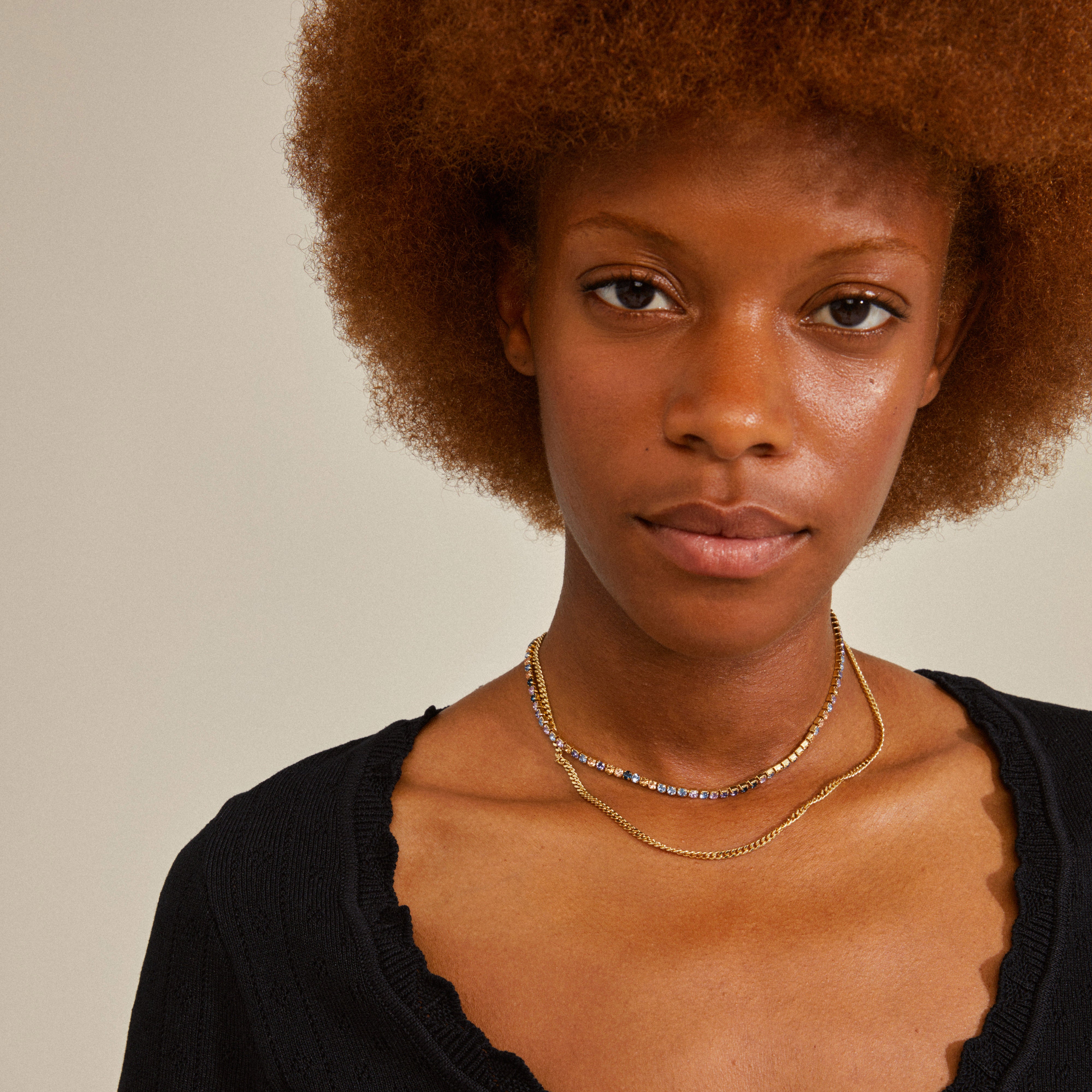 REIGN necklaces, 2-in-1 set, featuring a gold-plated curb chain and sparkling blue and champagne crystals, worn by a woman in a black shirt.