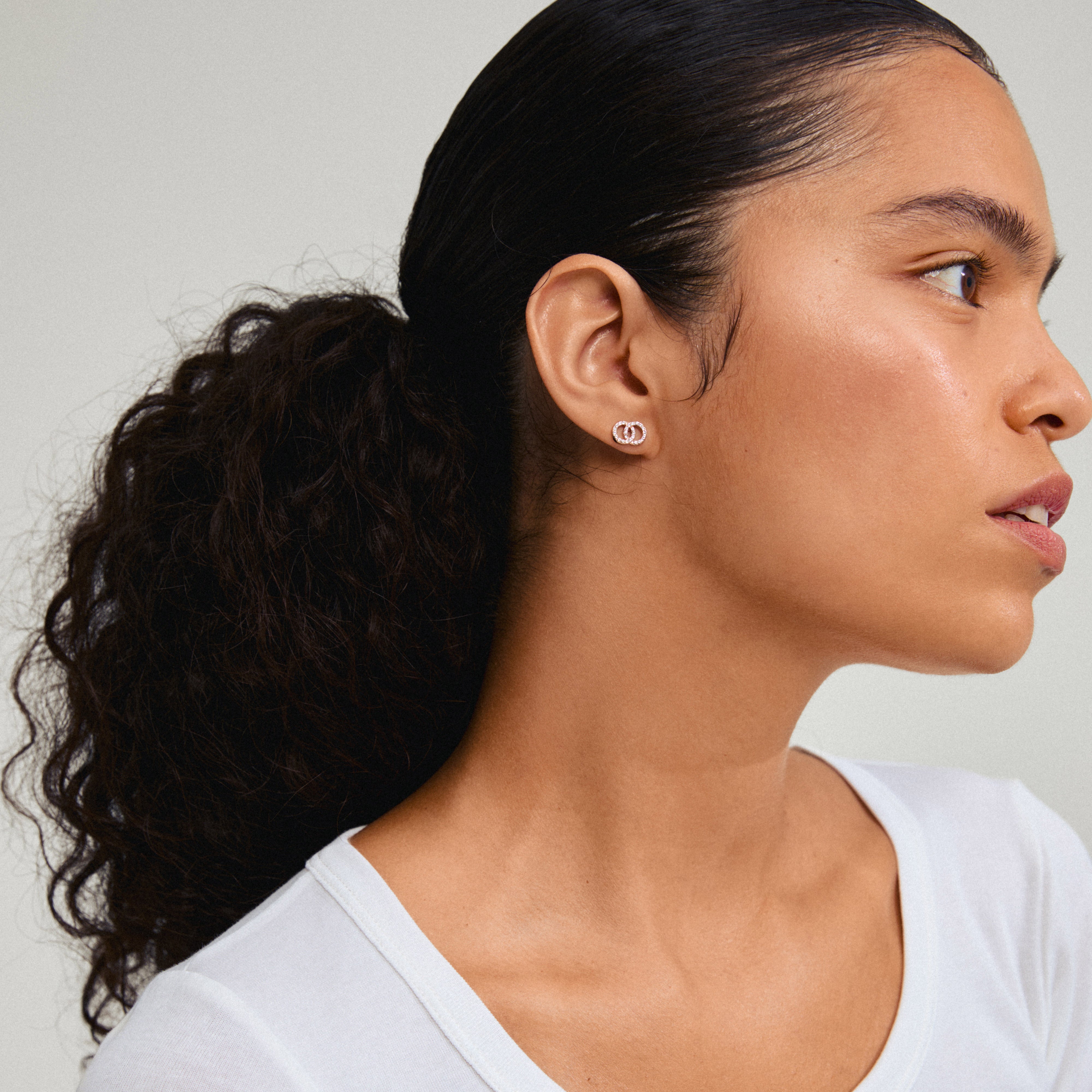 JOLINA recycled earrings in rose gold-plated design featuring intertwined halos with sparkling crystals, worn by a woman with a ponytail and visible ear.