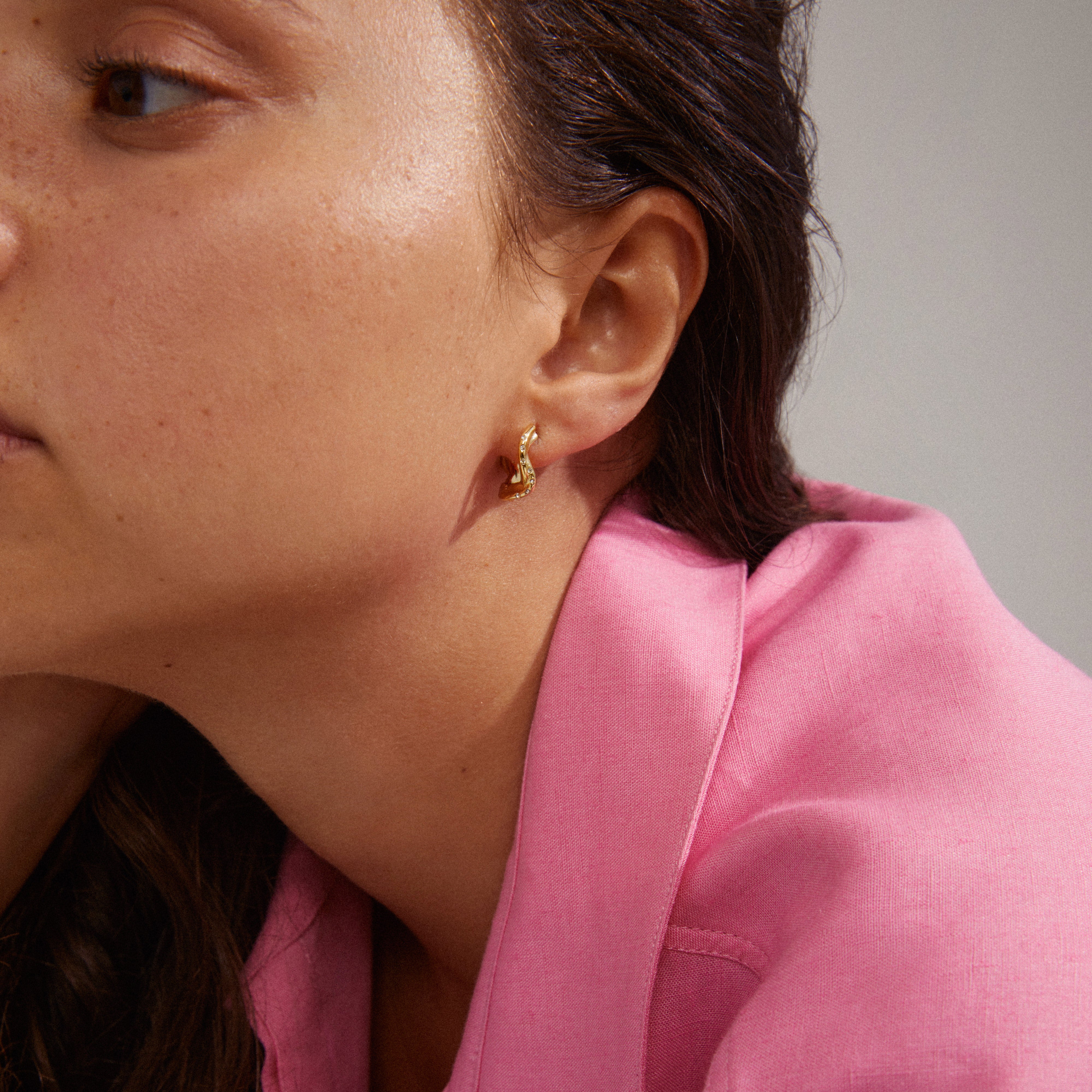 Close-up of a woman's ear adorned with Evra organic-shaped, gold-plated crystal hoops, showcasing their wavy design and discreet shine.