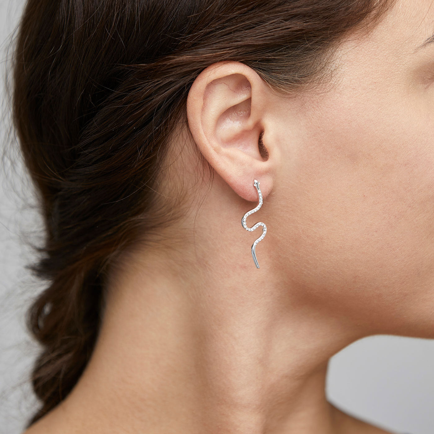 Close-up of a woman's ear wearing DAGNY crystal earrings, featuring silver-plated serpents adorned with sparkling crystals, coiling elegantly below the earlobes.
