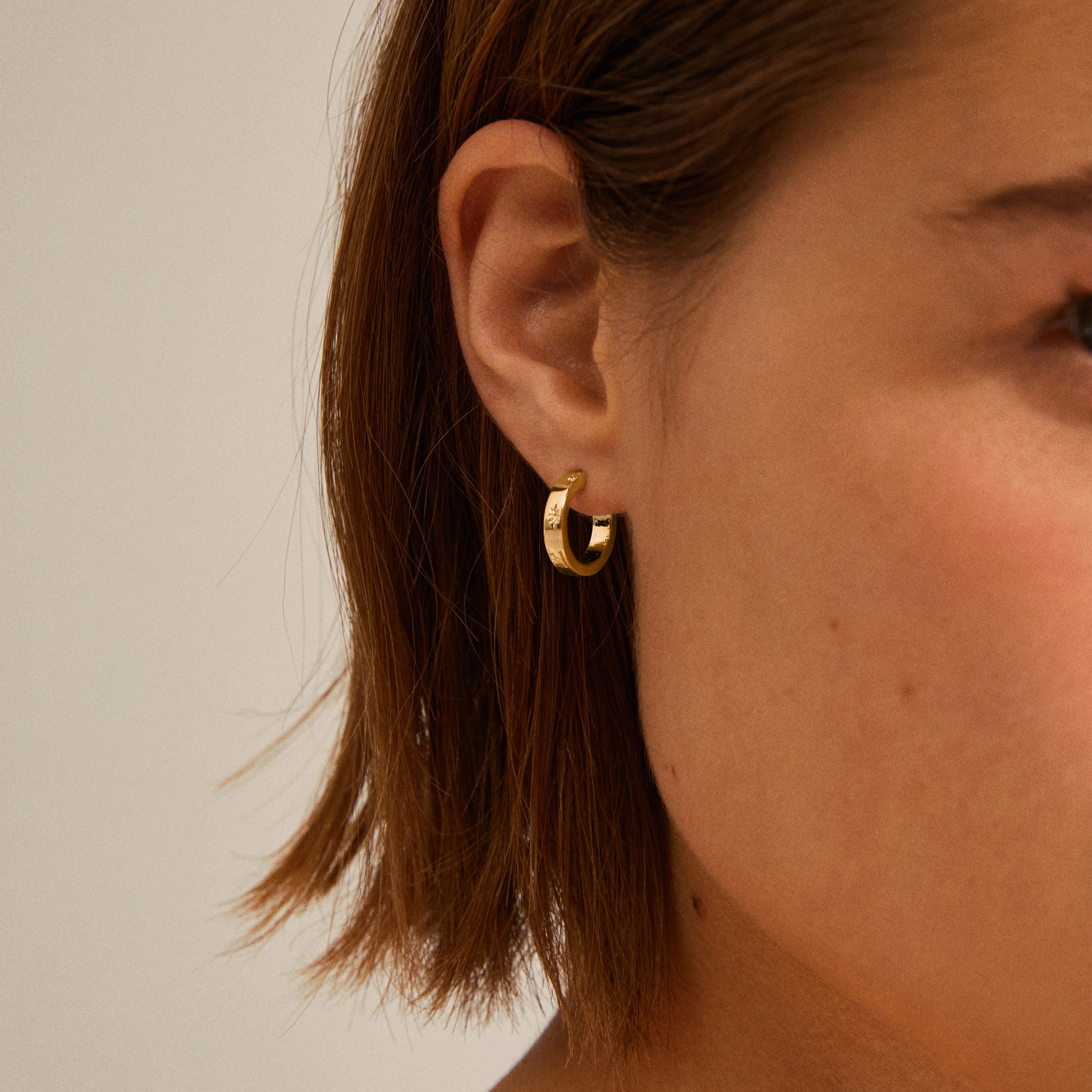 Close-up of a woman's ear wearing gold-plated Efia huggie hoop earrings with starry engravings, showcasing the elegant and dreamy design by Pilgrim.