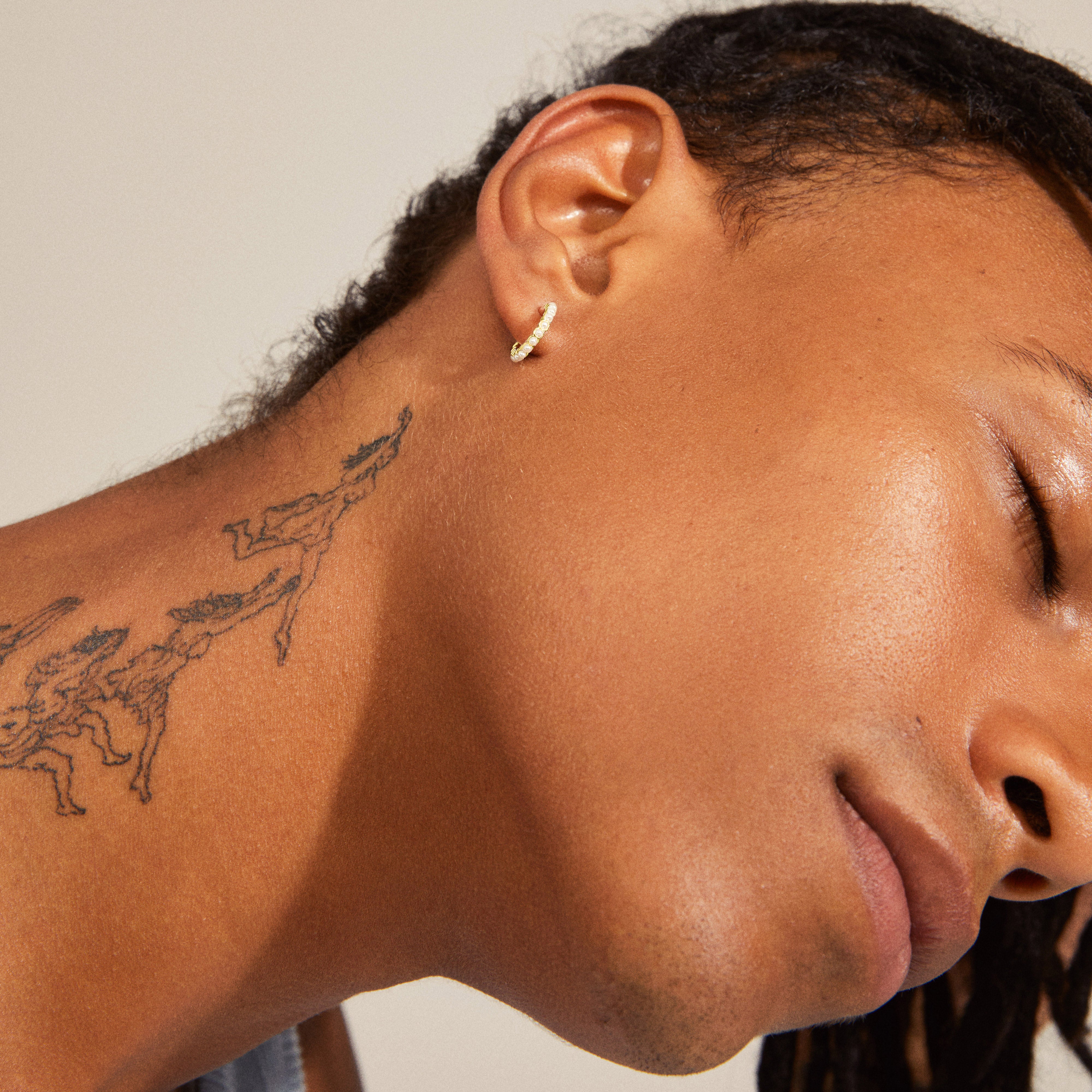 EKTA pearl huggie hoop earrings, gold-plated, featuring a row of pearls, worn by a person with a tattooed neck.