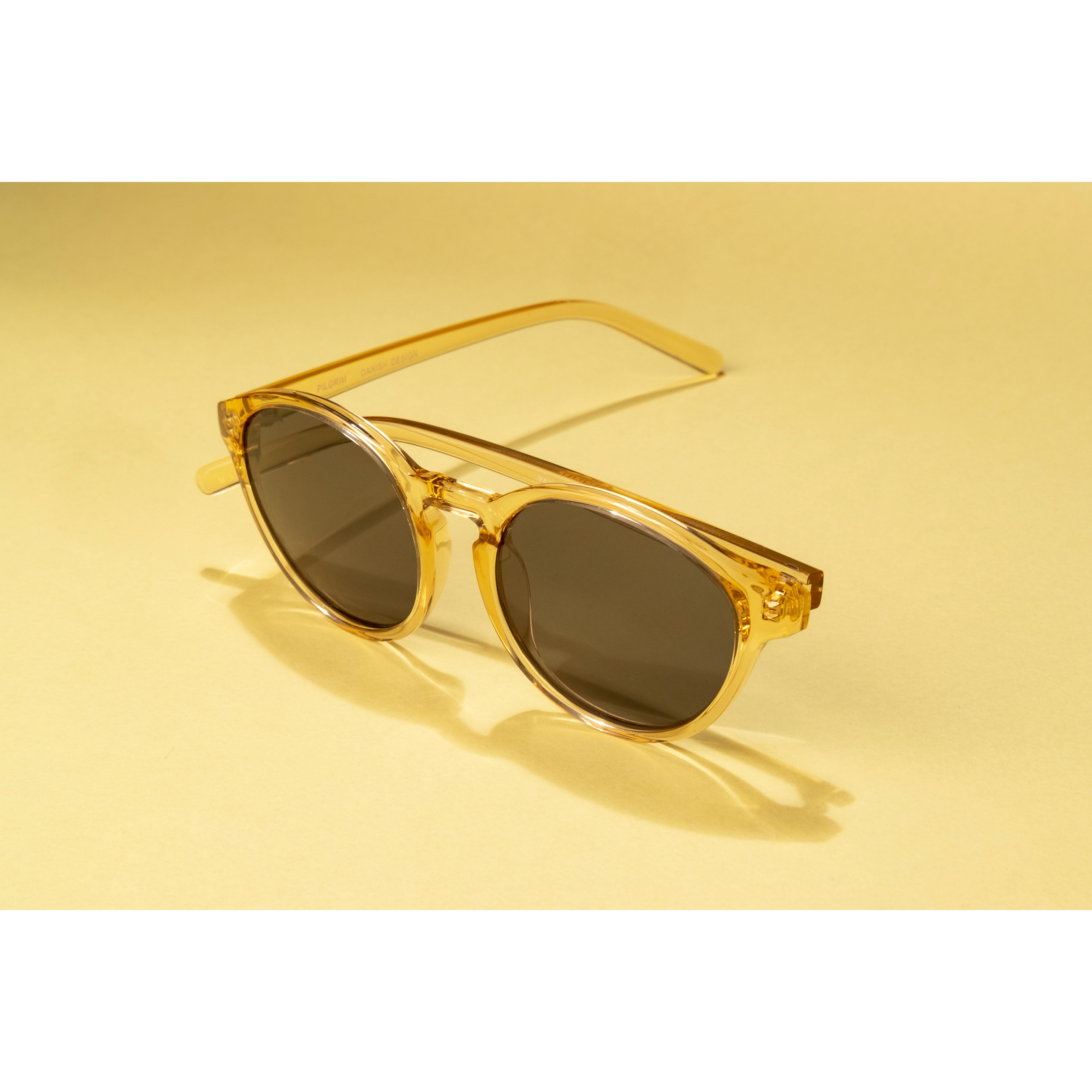 VASILIA sunglasses with a transparent frame and dark lenses on a yellow surface, embodying a trendy, feminine style with UVA/UVB protection.