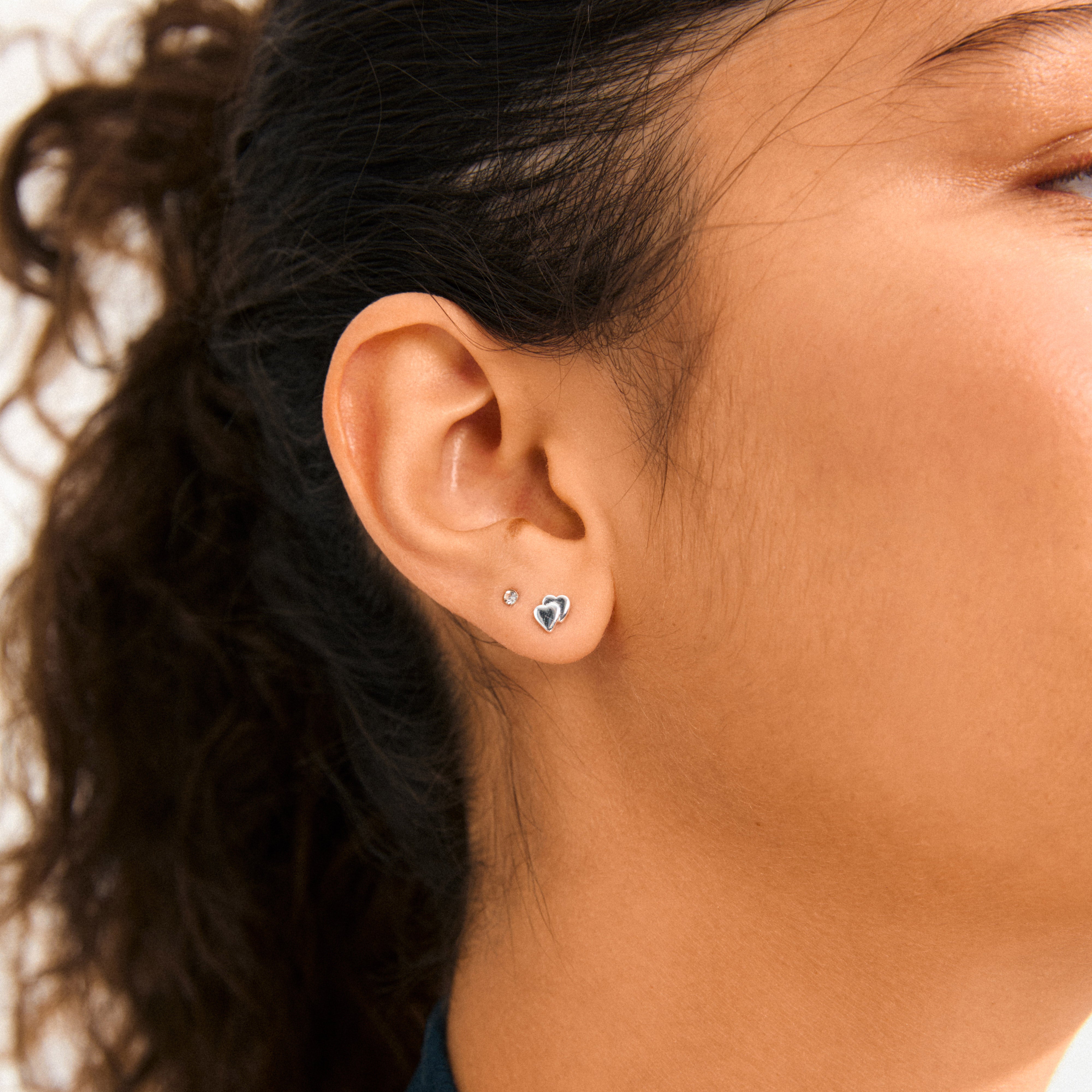 Close-up of a person's ear wearing HALO gift set earrings: a crystal stud and a silver heart stud, showcasing Pilgrim's trendy, eco-friendly Danish design.