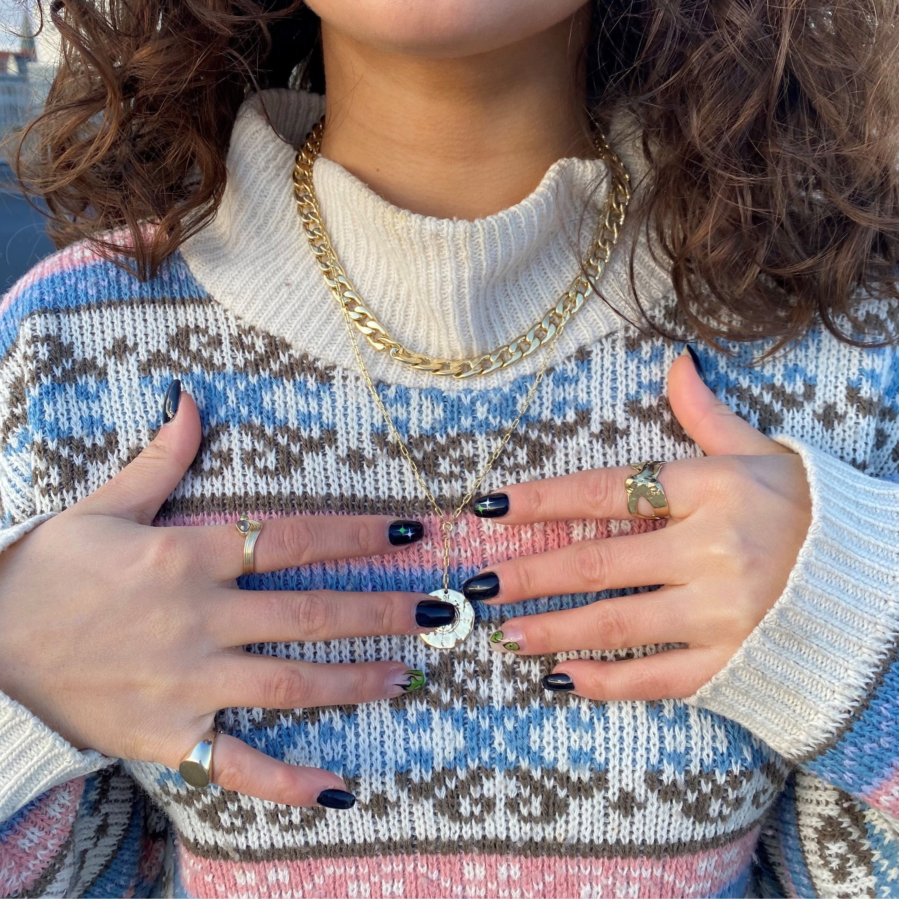 Person wearing a chunky sweater showcases the COMPASS organic-shaped, gold-plated ring with a rough, hammered texture, inspired by nature, alongside other gold jewelry.