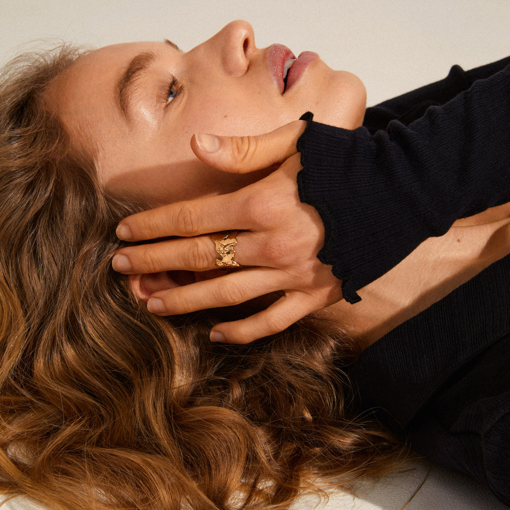 Person lying with light-brown hair, wearing a black ribbed sleeve, showcasing the COMPASS organic-shaped, gold-plated ring with a hammered texture, emphasizing natural, adventurous style.
